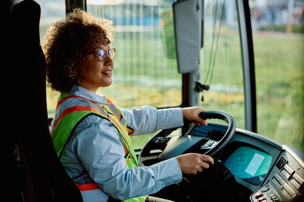 A woman with curly hair, glasses, and a safety vest driving a bus.