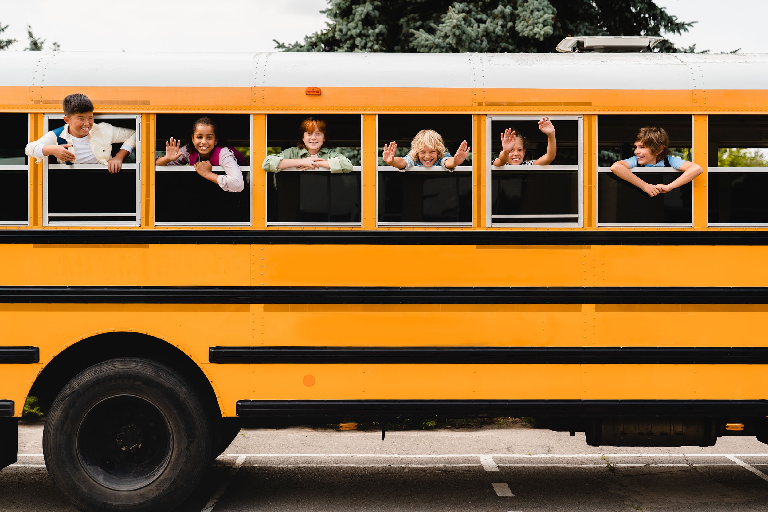 Children smiling and waving from the windows of a yellow school bus.