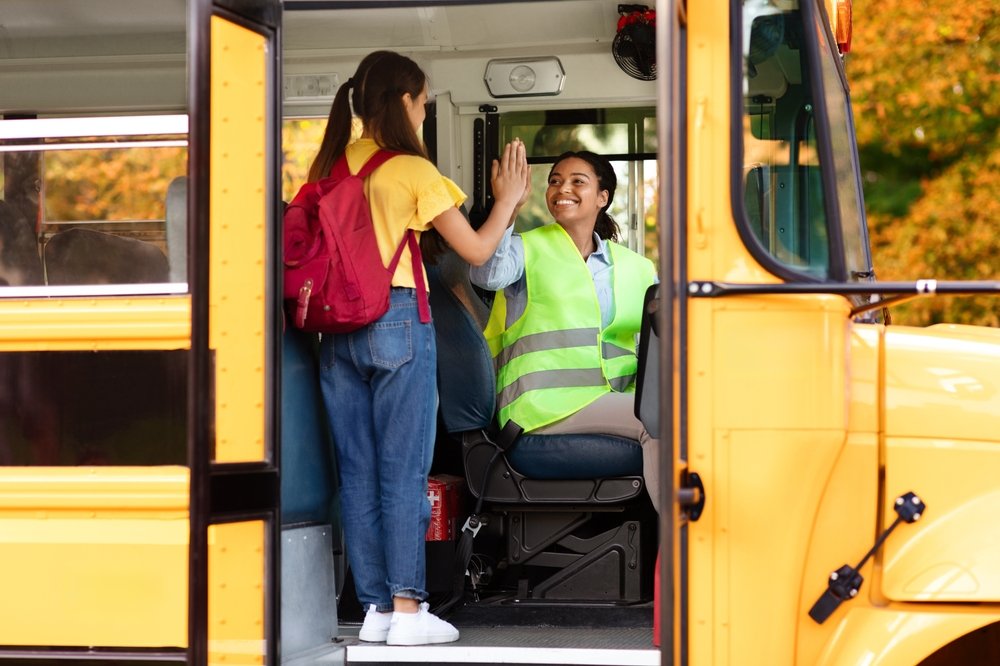 A young girl high-fives a bus driver inside a school bus, with fall foliage visible outside.