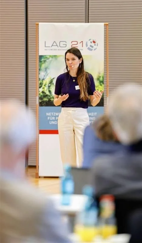 Stella Schaller. Eine junge Frau steht bei einer Präsentation, im Hintergrund ein Stand mit der Beschriftung "LAG21" und einem Logo. Sie hält ein Handy in der Hand und spricht vor einem Publikum in einem Raum mit Holzlaminat.