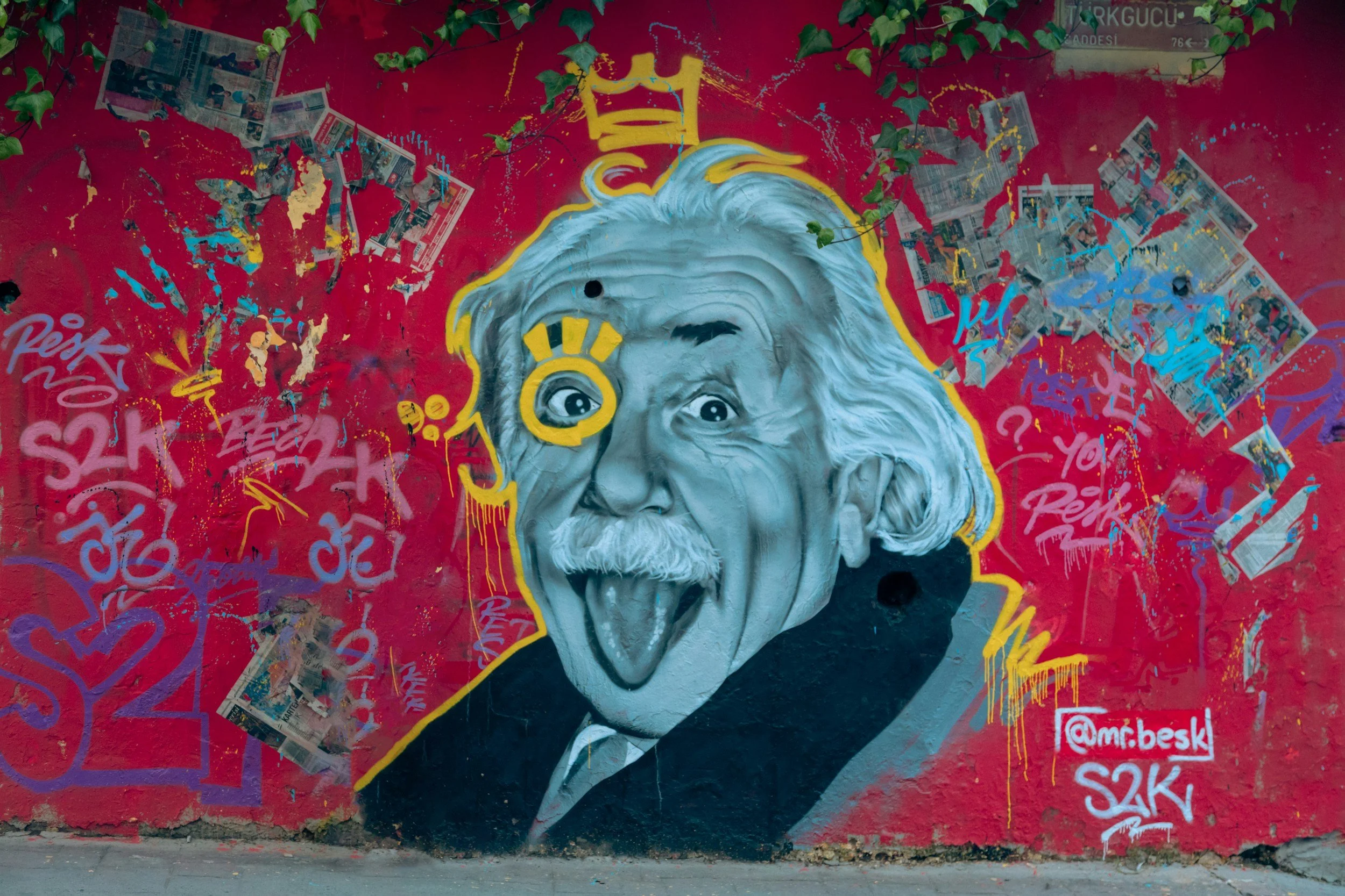 Graffiti-Mural eines alten Mannes mit langem weißen Haar und Bart, auf rotem Hintergrund, mit verschiedenen Skizzen und Aufklebern drum herum.