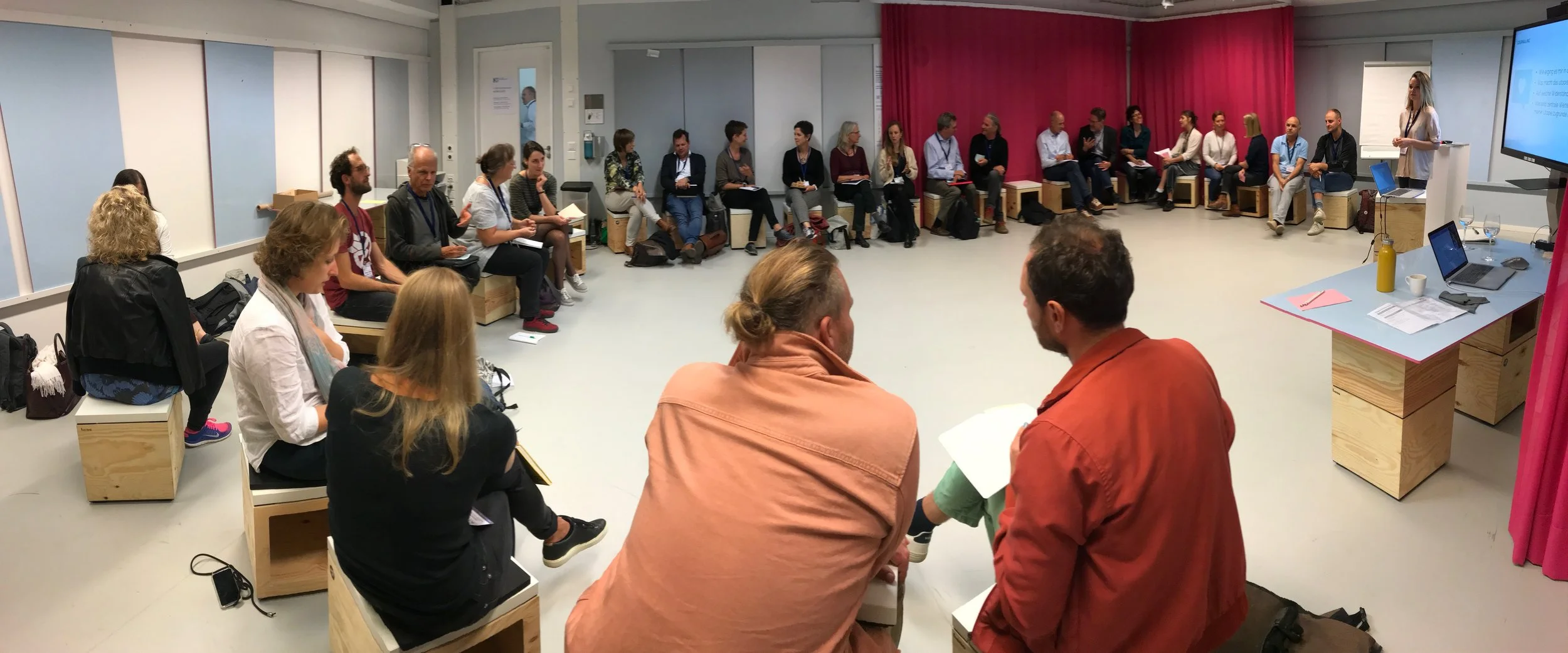 Neunundzwanzig Personen sitzen im Kreis in einem Raum, die meisten mit Notizbüchern oder Papieren in der Hand, während eine Frau an der Front eine Präsentation hält. Es gibt einen Tisch mit einem Laptop, Wasser, Notizblöcken und anderen Gegenständen. Der Raum hat graue Wände, eine rote Vorhangwand im Hintergrund und eine Leinwand oder Bildschirm an der rechten Seite.