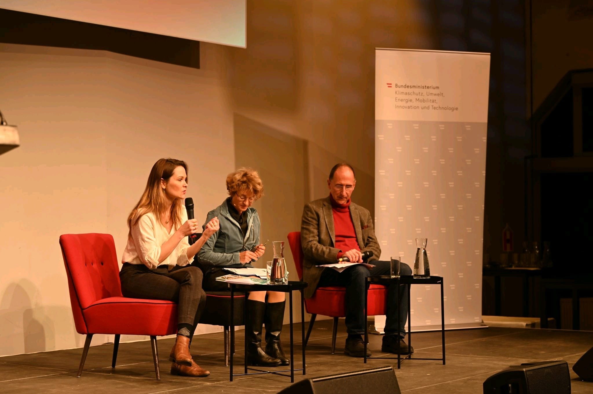 Stella Schaller - Panel.Menschen sitzen auf einer Bühne bei einer Diskussion, mit einem Mikrofon, Wasserflaschen und Unterlagen. Im Hintergrund ein Banner des Bundesministeriums für Klimaschutz, Umwelt, Energie, Mobilität, Innovation und Technologie.