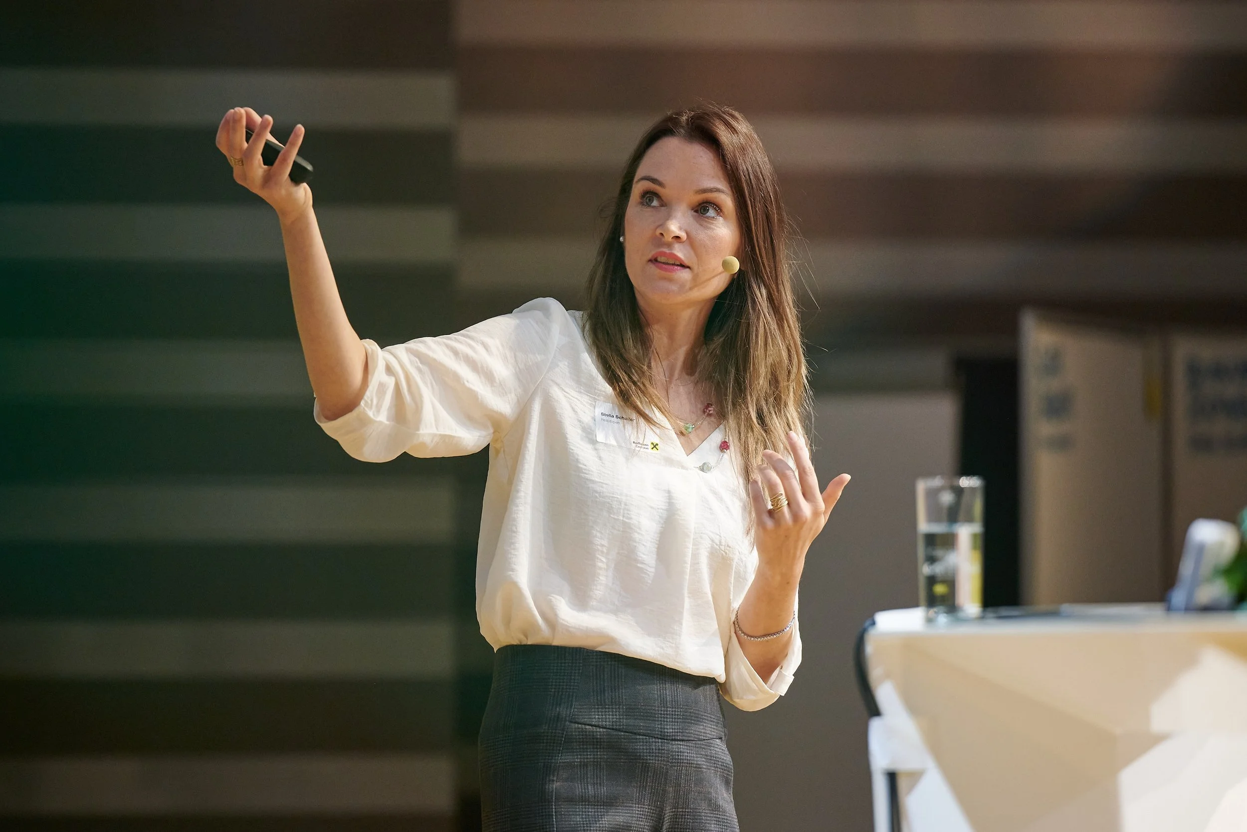 Stella Schaller. Eine Frau mit braunen Haaren, die eine weiße Bluse trägt, hält eine Fernbedienung in der Hand und spricht während eines Vortrags in einem Raum mit dunklem Hintergrund.