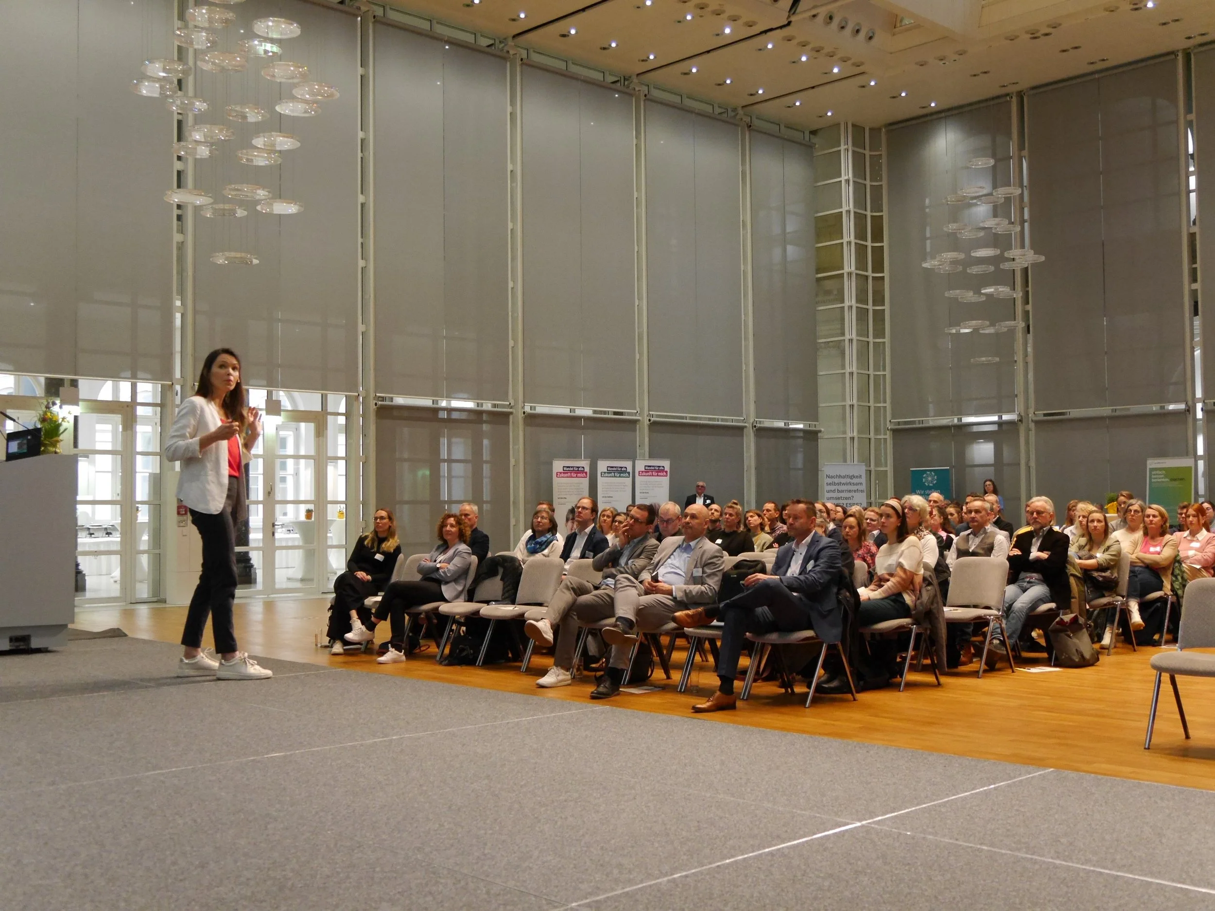Stella Schaller. Ein Vortragspodium mit einer Frau, die vor einem Publikum spricht. Das Publikum sitzt in Stühlen und hört aufmerksam zu, im Hintergrund zahlreiche Banner und Plakate, hoher Saal mit Deckenbeleuchtung.