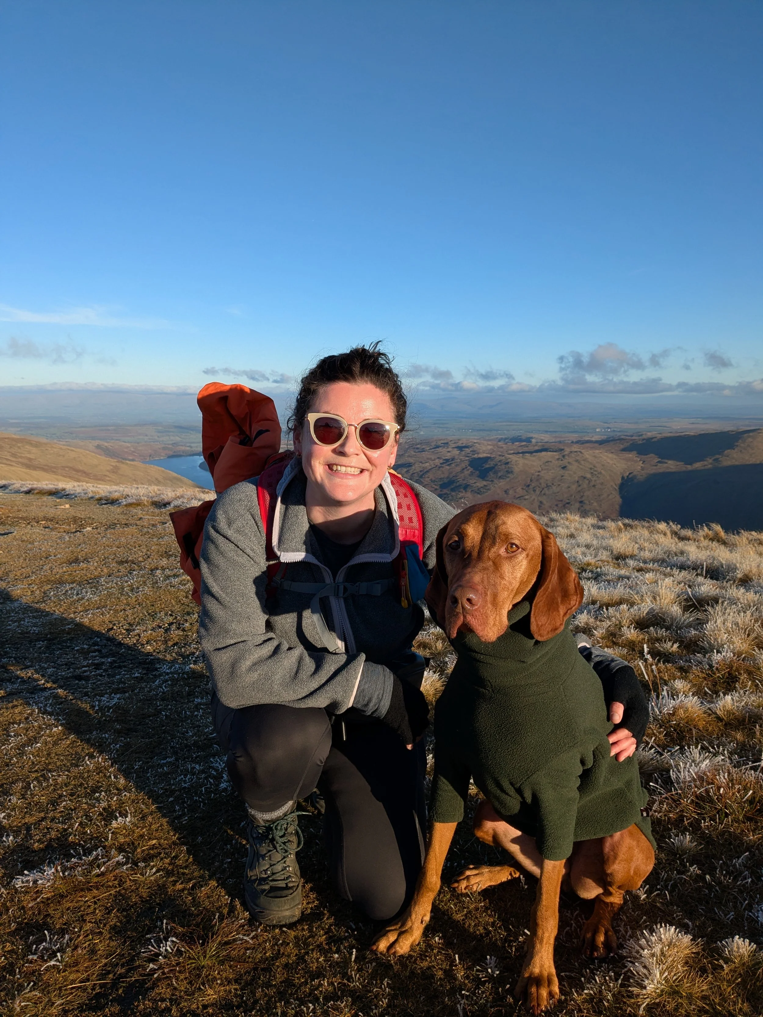 Zara and her vizsla Jeffrey smile for the camera on a mountain hike with a scenic view of hills and a clear blue sky in the background.