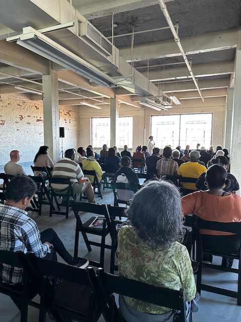 People seated in chairs attending a presentation or lecture in an industrial-style room with brick walls and large windows.