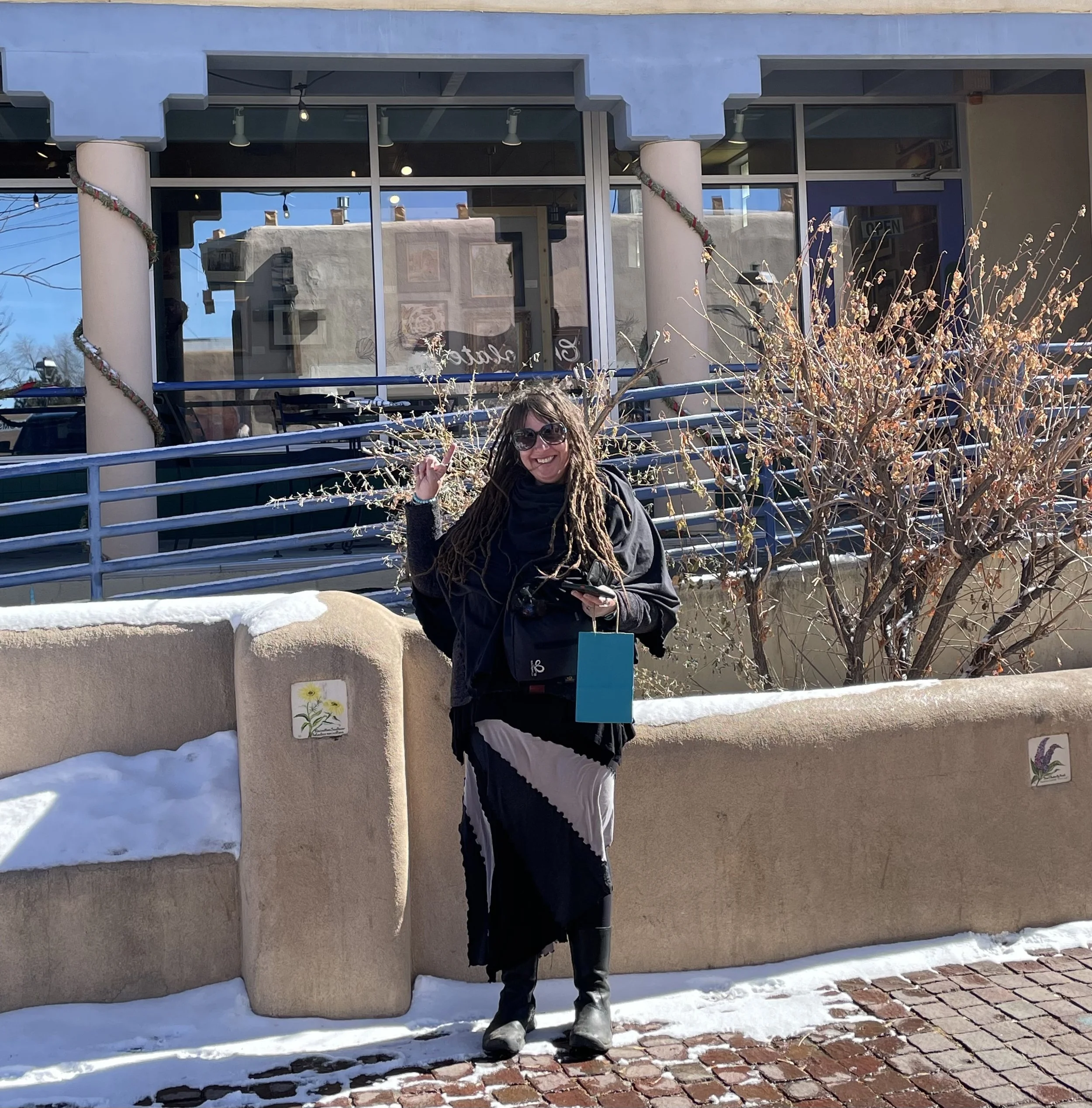 A woman with long dreadlocks wearing sunglasses and black jacket smiling and pointing, standing outdoors in front of a building with large glass windows and beige walls. Snow is on the ground and on a decorative wall with flowers painted on it, and there are dry bushes in the foreground.