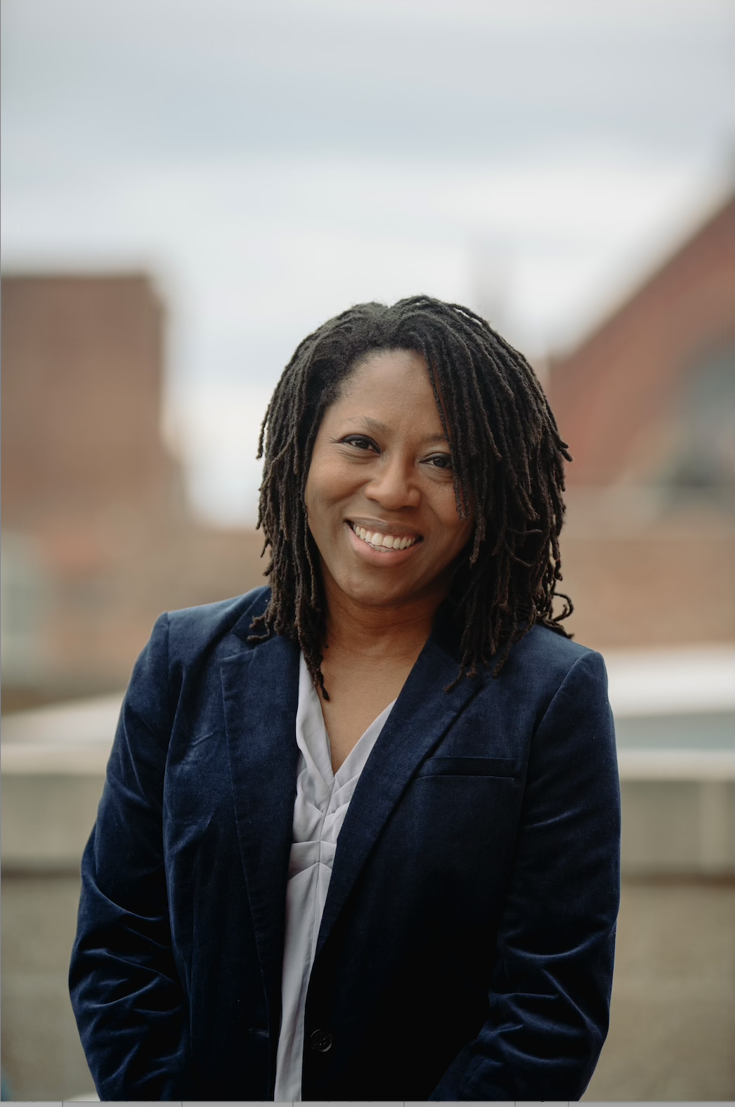 A smiling woman with shoulder-length dreadlocks wearing a navy blazer and white blouse outdoors with blurred background of buildings.