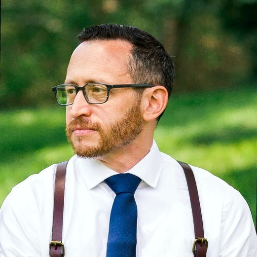 A man with glasses, a beard, wearing a white shirt, blue tie, and suspenders outdoors with a green blurred background.