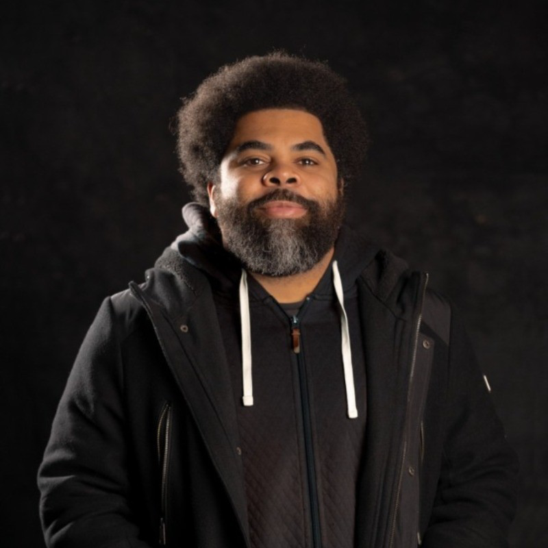 A man with a full beard and afro hairstyle wearing a black jacket and hoodie, standing against a dark background.