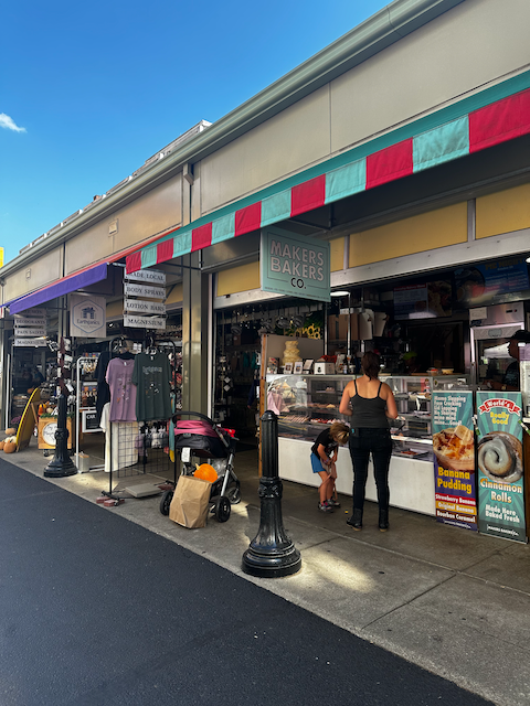 A woman and a young girl are standing at an ice cream shop called Makers & Bakers Co. in a shopping area. The shop has a colorful striped awning, and there are signs advertising banana pudding and cinnamon rolls. Outside, clothing and other items are displayed for sale, and there is a stroller with a bag next to it.