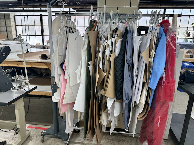 A clothing rack filled with fabric samples and material swatches in various colors and textures, in a well-lit sewing or fashion design studio.