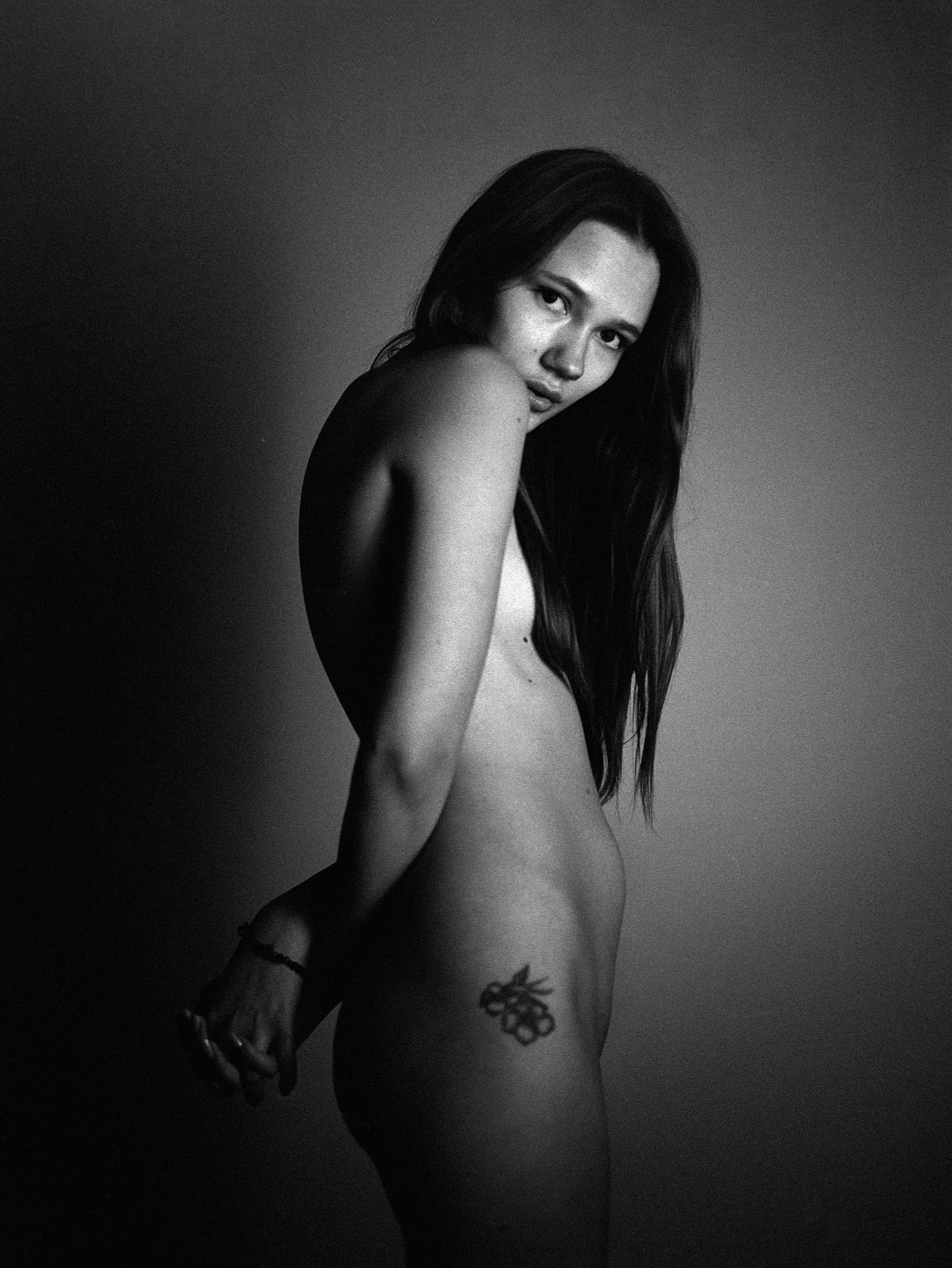 Schwarz-Weiß-Foto eines nackten Frauenkörpers mit langem Haar, der zur Kamera schaut, mit Tätowierung am Oberschenkel.