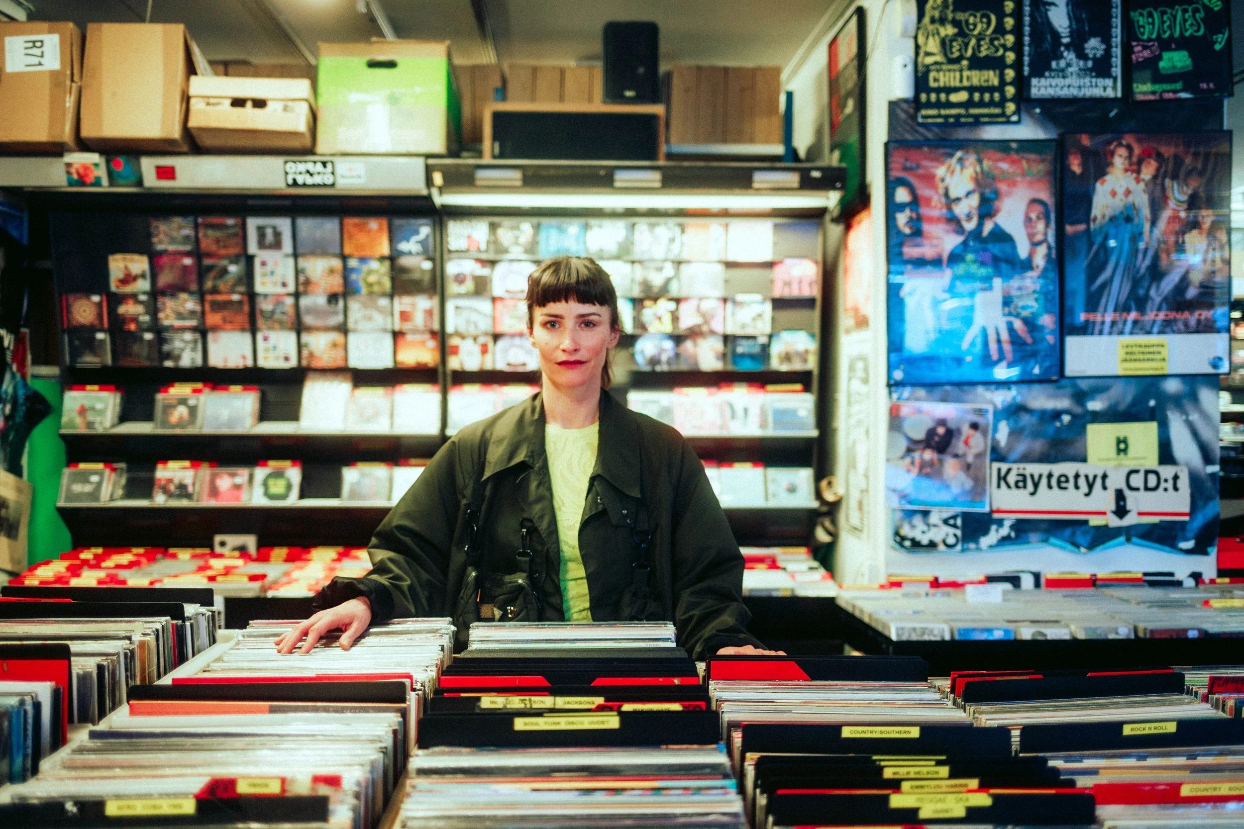 Eine junge Frau steht in einem Plattenladen, umgeben von Vinyl-Schallplatten, die in Regalen und auf Tischen angeordnet sind.