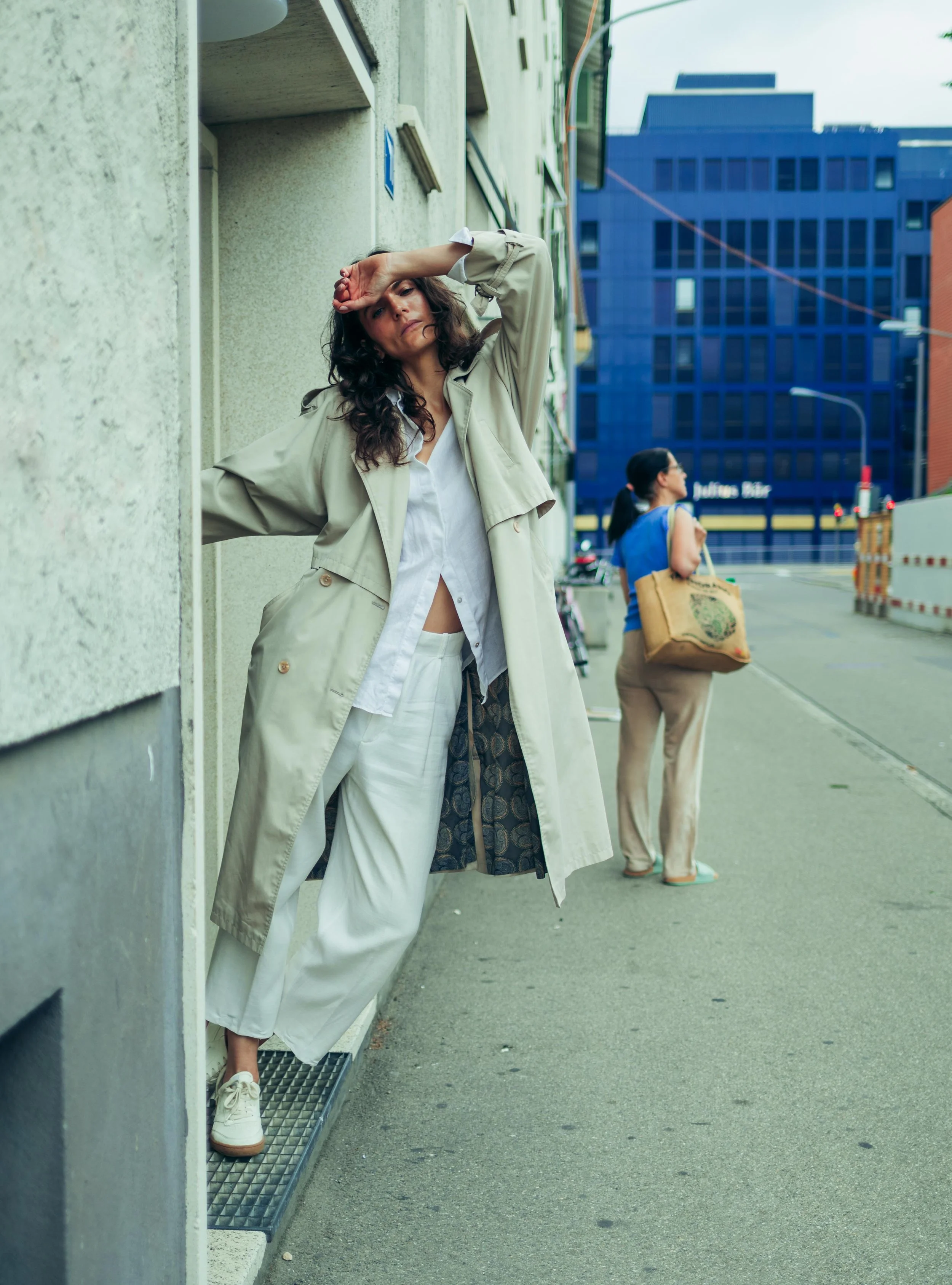 Eine Frau lehnt an einer Hauswand, trägt eine beige Trenchcoat, weiße Hose und Sneakers, wirkt müde oder nachdenklich. Im Hintergrund steht eine andere Frau mit Einkaufstasche, urbanes Stadtbild mit modernen Gebäuden.