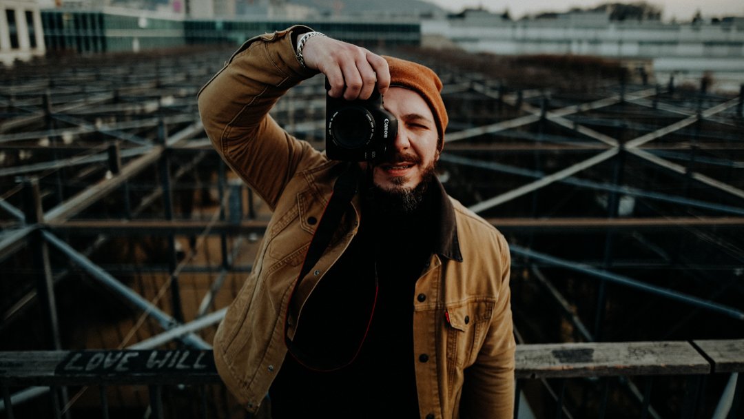Mann mit braunem Hut und Jacke, der ein Kamera fotografiert, steht auf einem Dach mit einer Metallstruktur im Hintergrund.