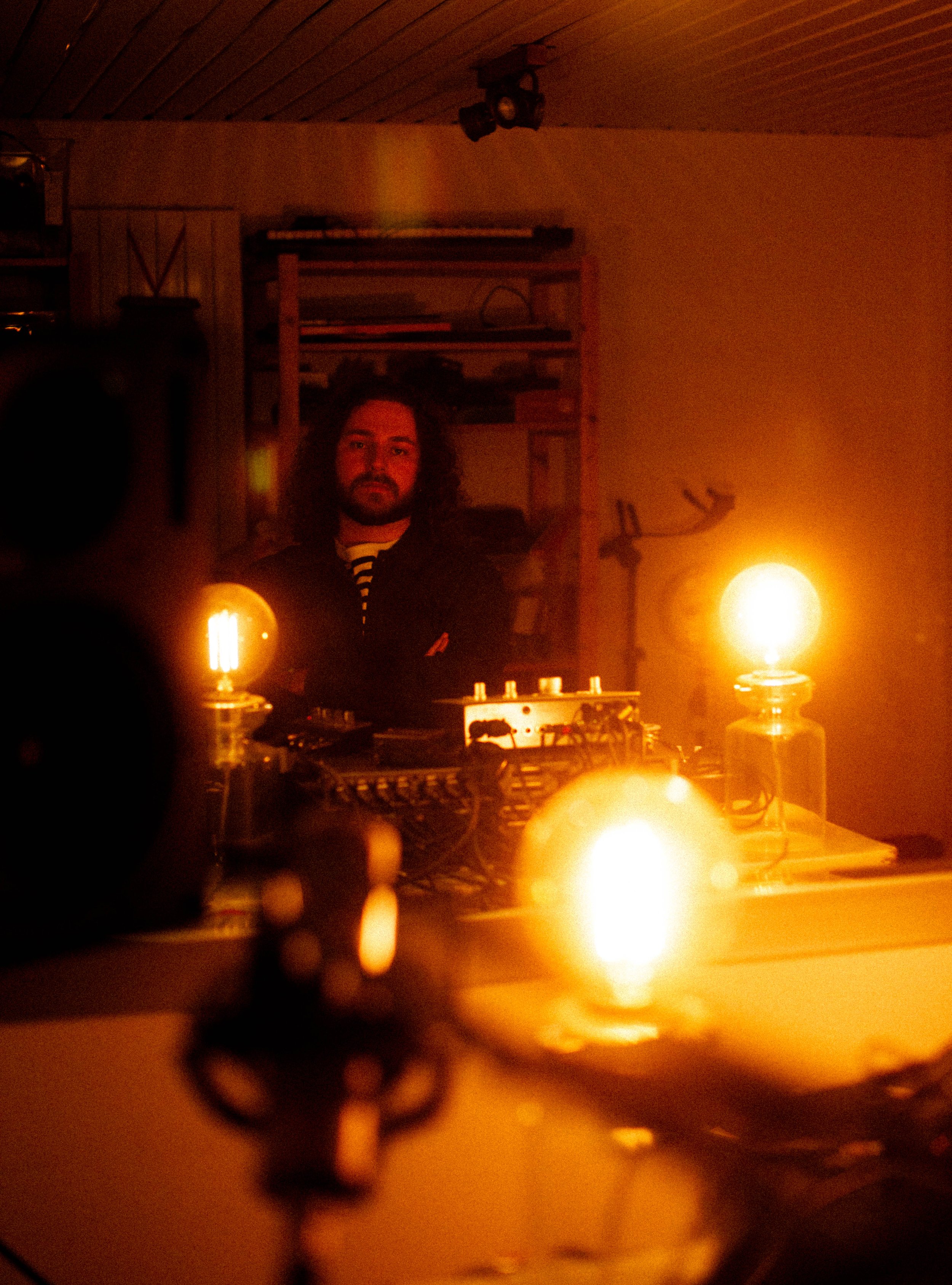 Ein Mann mit langen Haaren steht in einem Raum mit warmem Licht, umgeben von Musikequipment und zwei sichtbaren Glühbirnen, die für Stimmung sorgen.