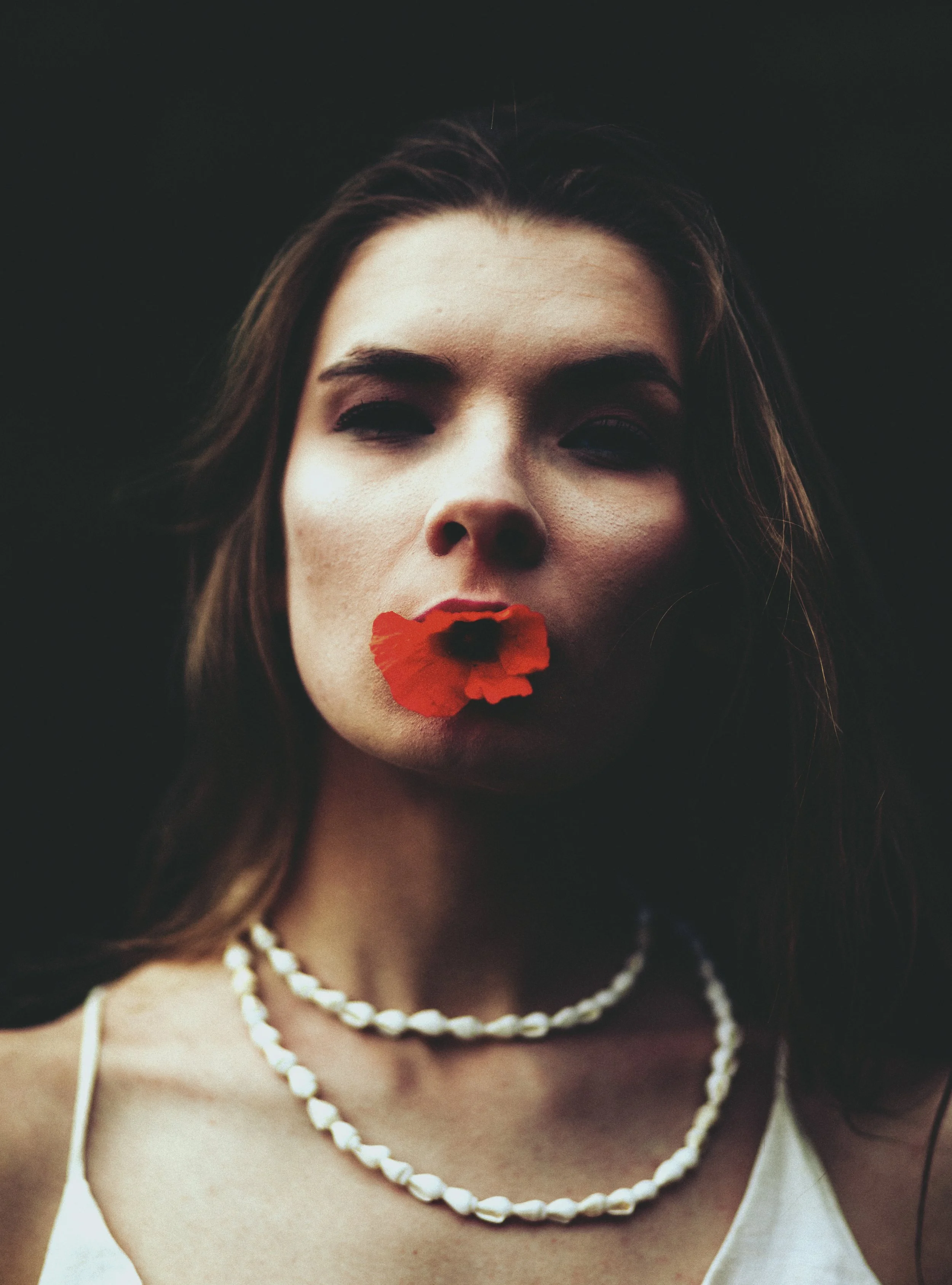 Porträt einer Frau mit einem roten Blütenblatt im Mund, schwarze Kleidung, dunkler Hintergrund.