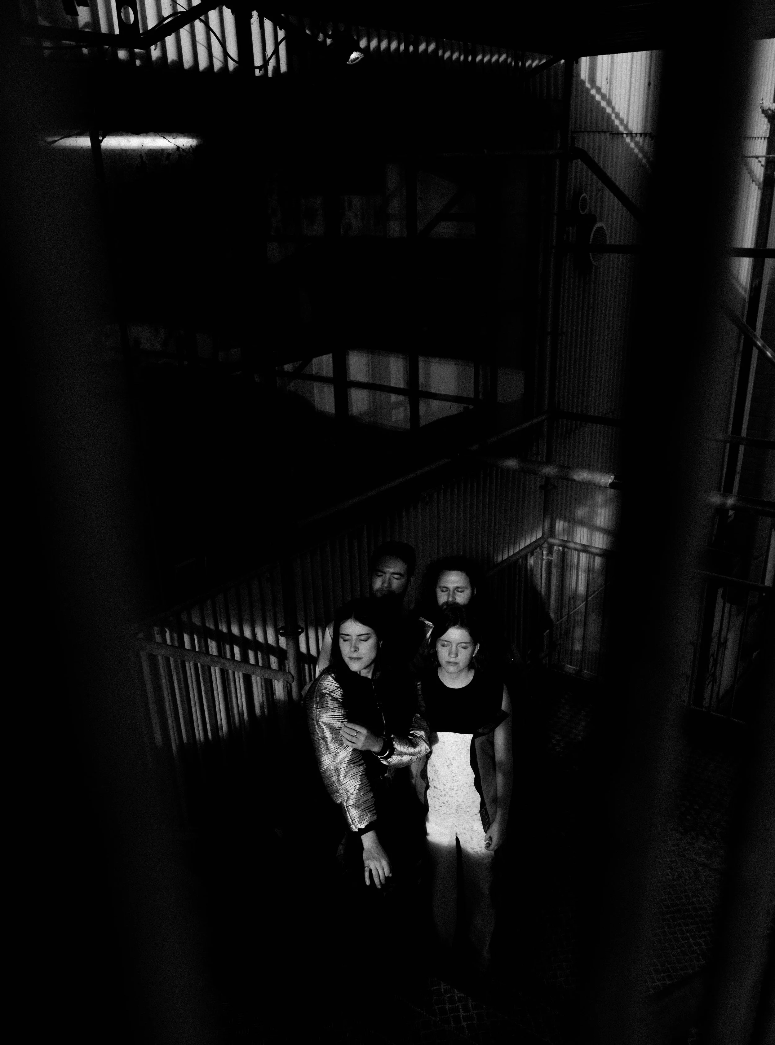 Fünf Menschen stehen in einem dunklen Treppenhaus, umgeben von Metallgeländern und -wänden, mit gedämpftem Licht. Sie stehen in einer Gruppe, einige mit verschränkten Armen, alle mit geschlossenen Augen.