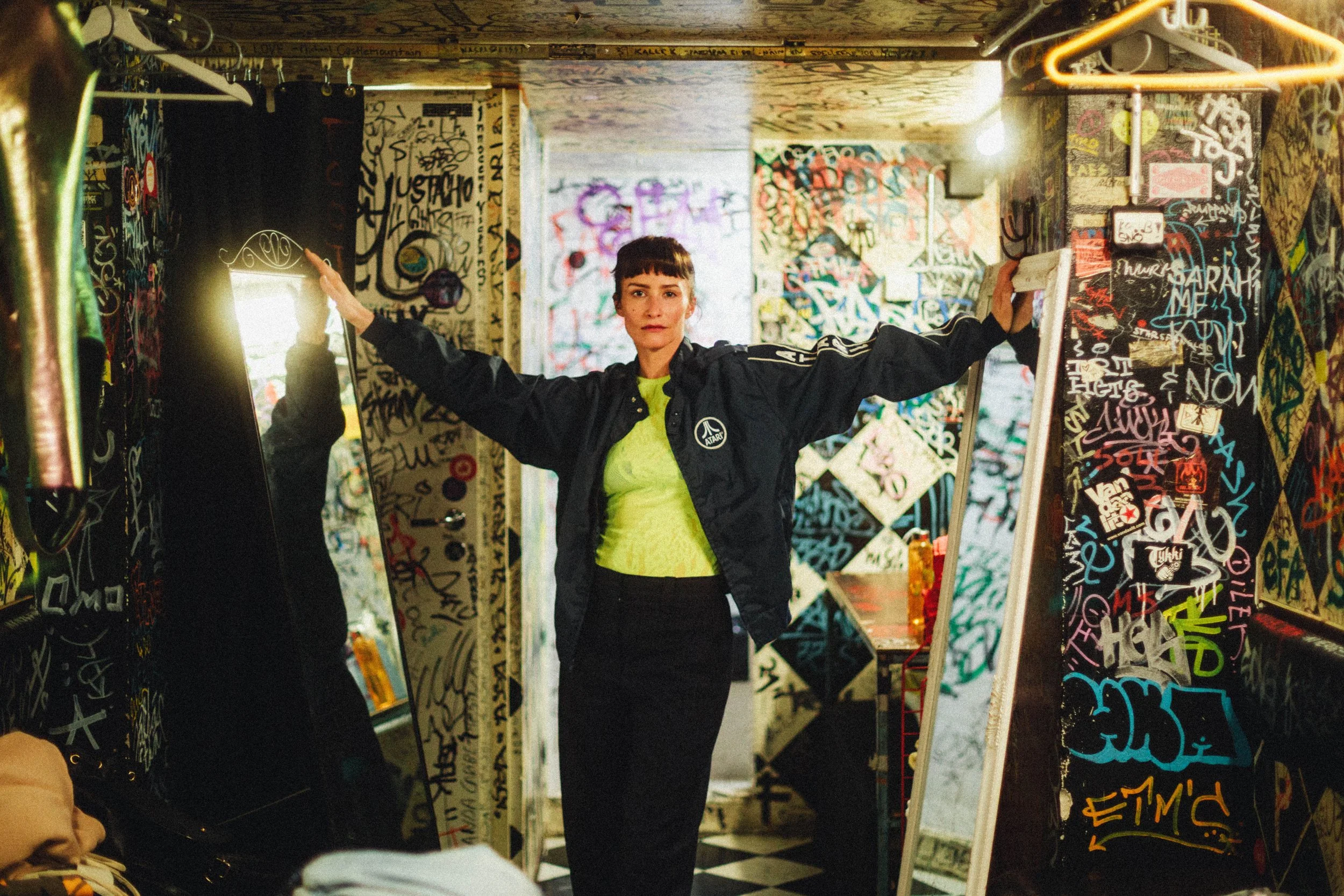 Frau steht in einem graffiti-verwachsenen Raum mit offenen Armen, trägt eine schwarze Jacke und ein neon grünes Oberteil, im Hintergrund bunte Graffiti-Wände.