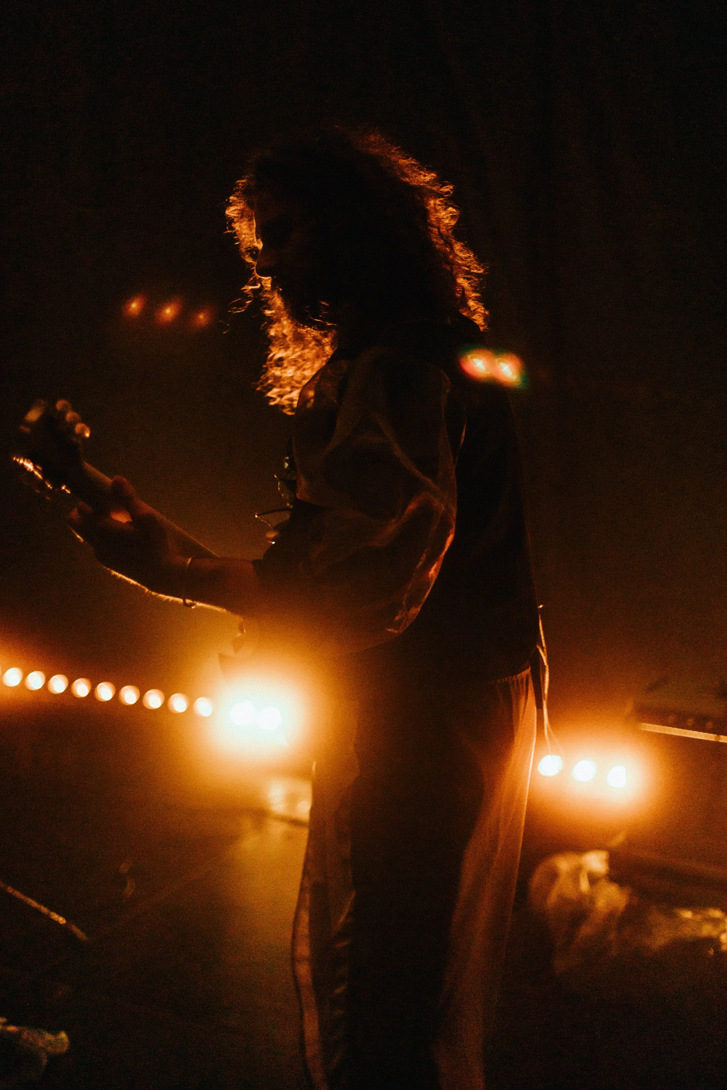 Eine Person mit lockigem Haar spielt Gitarre in einem dunklen Raum, beleuchtet von warmen Scheinwerfern im Hintergrund.
