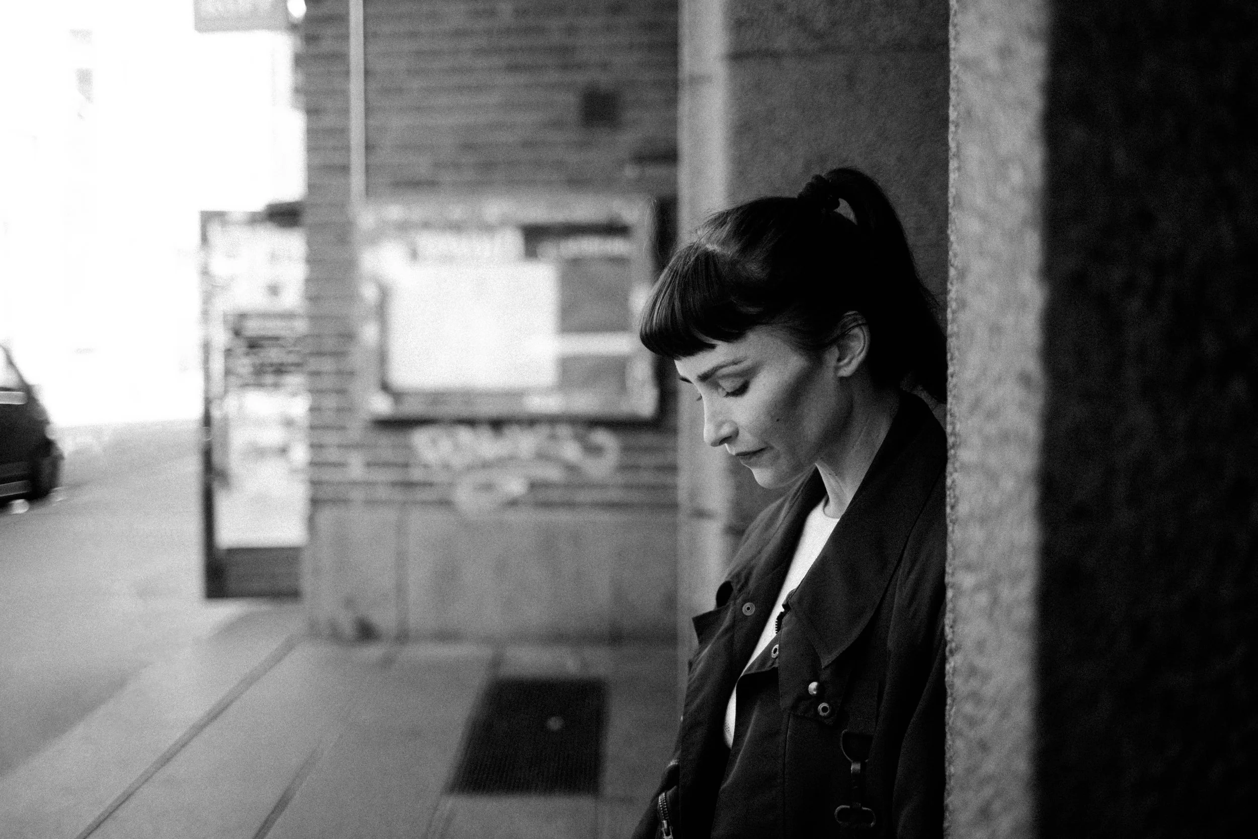 Schwarz-weiß-Foto einer Frau mit dunklen Haaren, die an einer Steinmauer an einer urbanen Straße lehnt, mit Blick nach unten und einem nach oben gebundenen Pferdeschwanz.