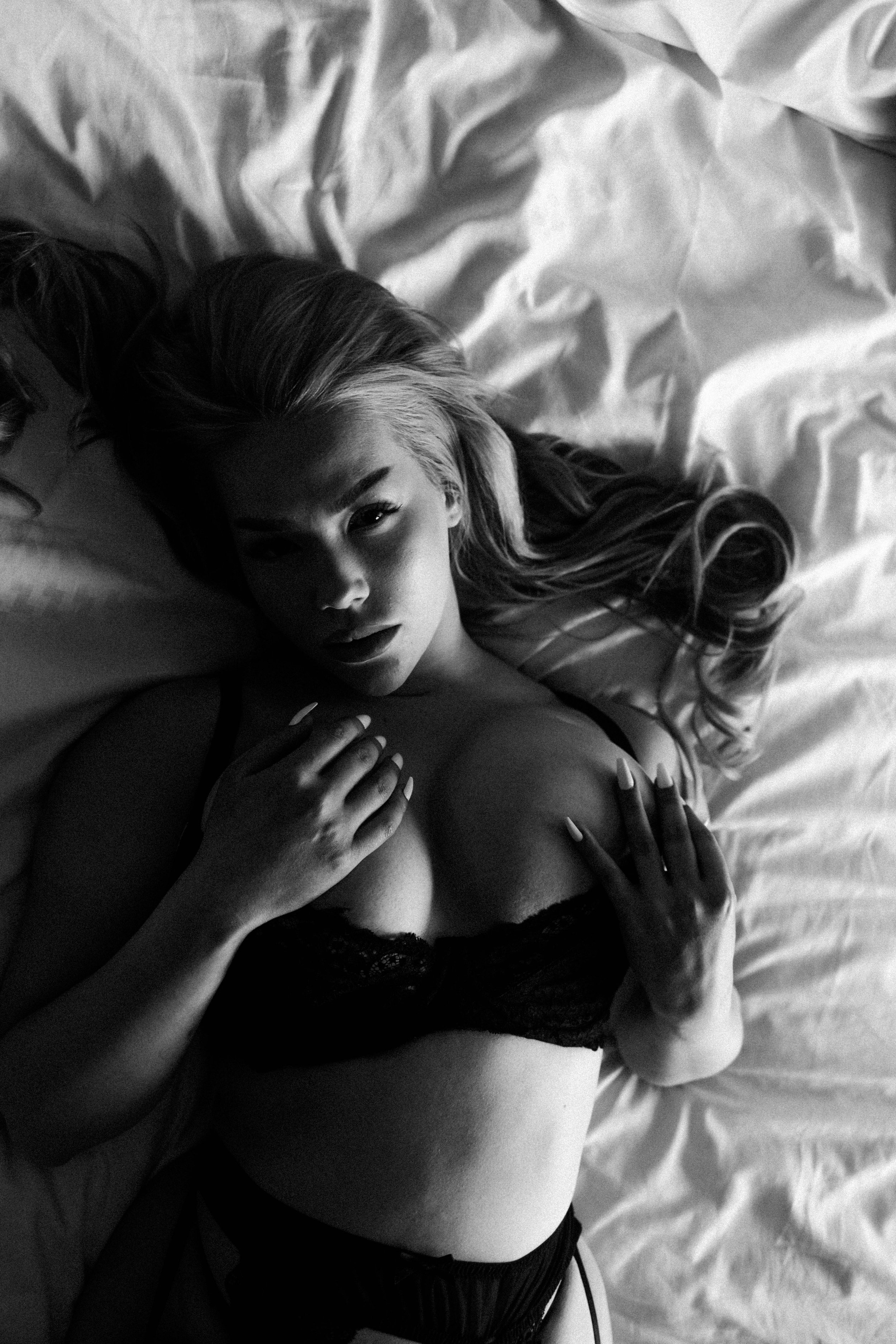 Schwarze und weiße Nahaufnahme eines liegenden Mädchens mit langen Haaren, in schwarzer Spitzeunterwäsche, blickt in die Kamera, auf einem Bett.