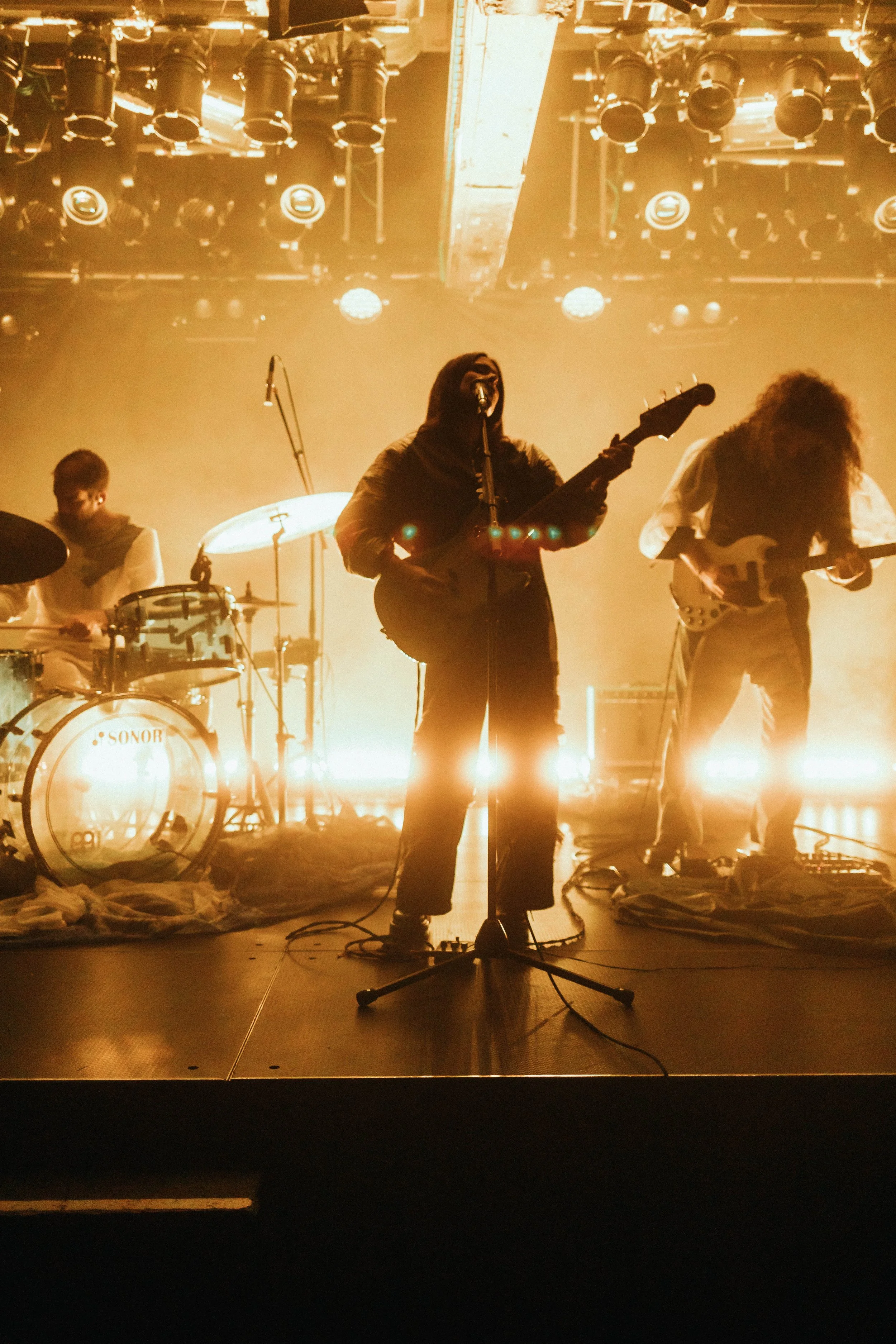 Band auf der Bühne während eines Konzerts, zurückhaltende Beleuchtung, Musiker spielen Gitarre und Schlagzeug, im Hintergrund Scheinwerfer und Bühnenlichter.