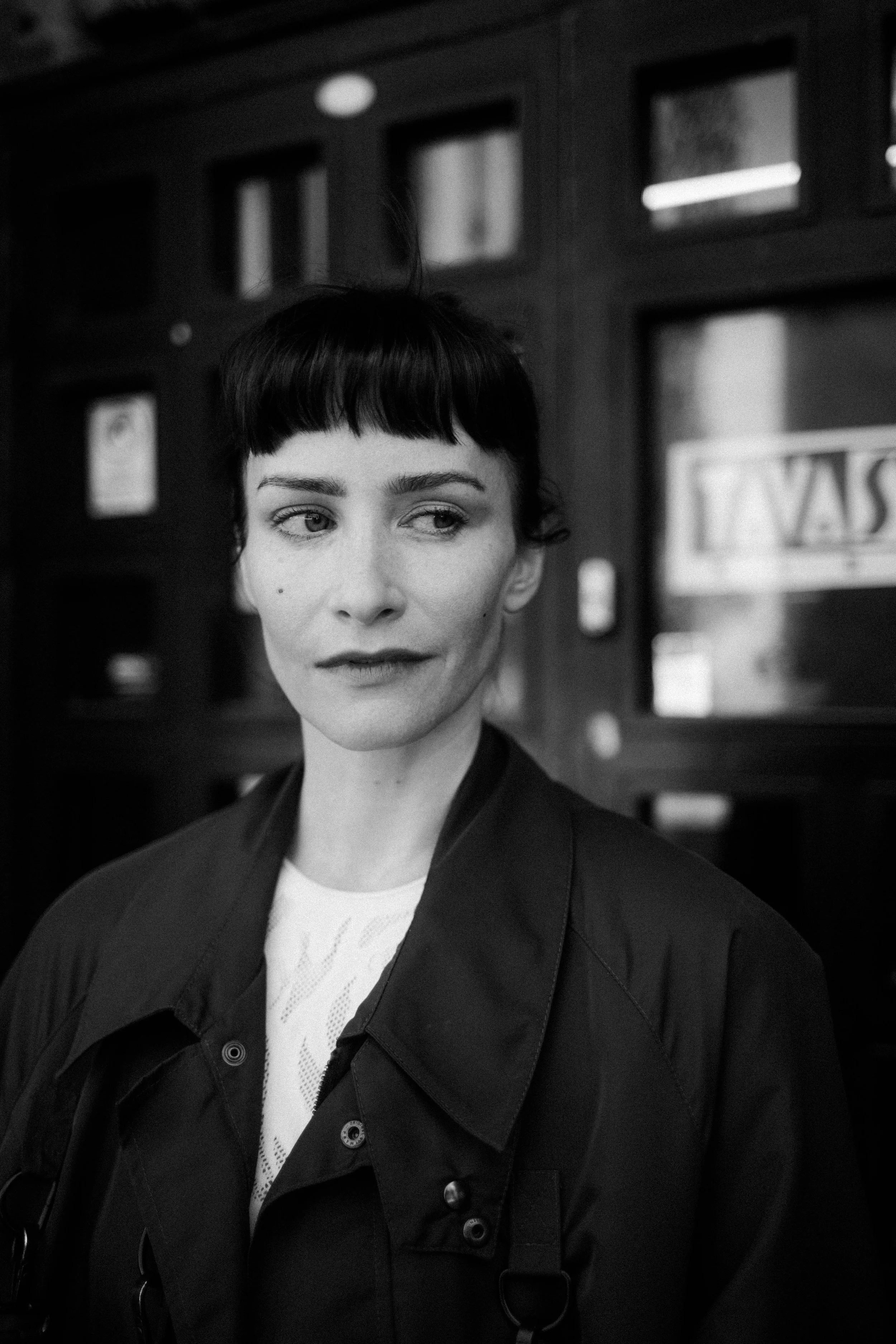 Schwarz-Weiß-Fotografie einer Frau mit dunklem Kurzhaarschnitt, die nach rechts blickt, vor einer dunklen Wand mit Fenstern