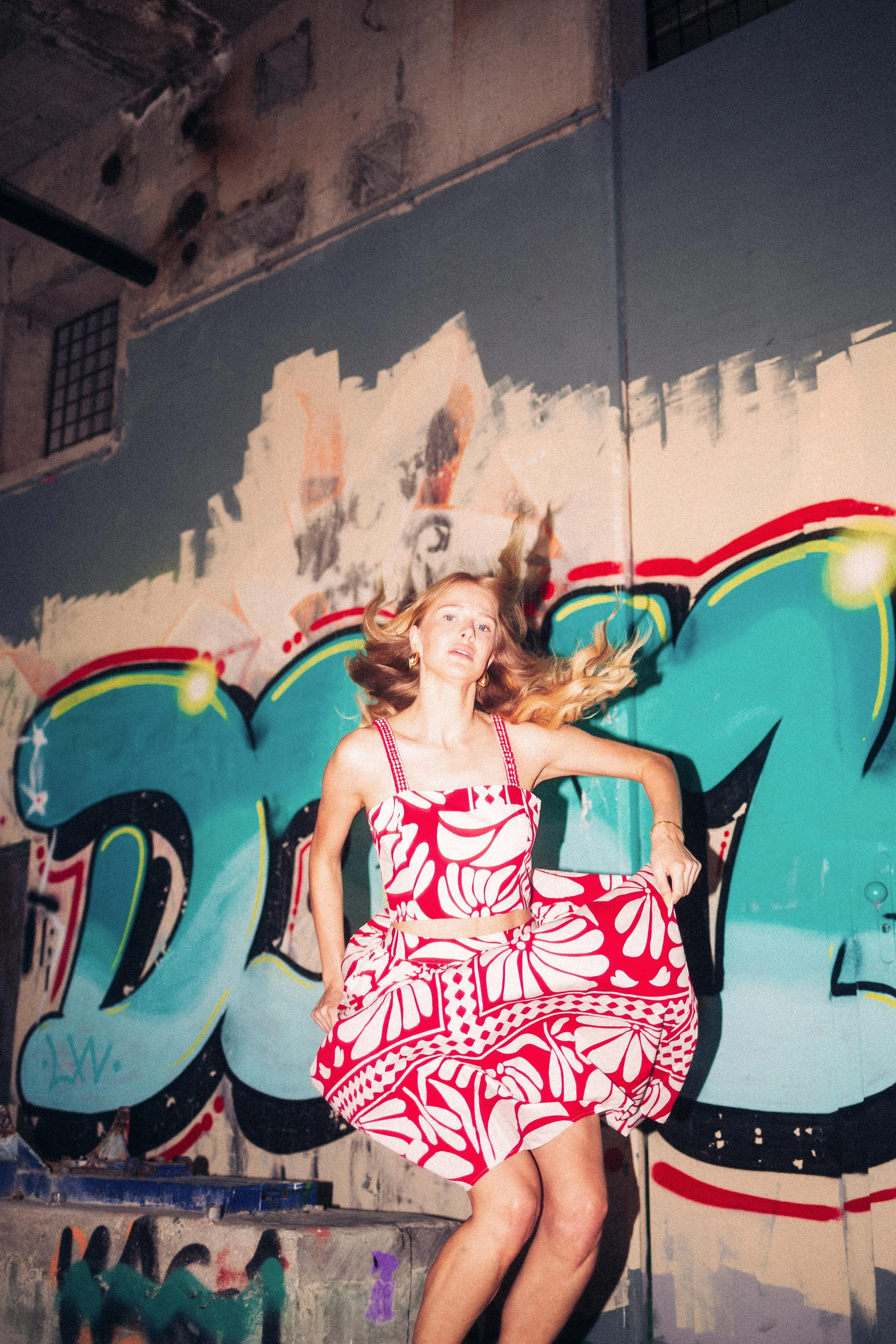 Frau in einem roten und weißen, floral gemusterten Kleid tanzt vor einer bunten Graffiti-Wand in einem urbanen Umfeld.