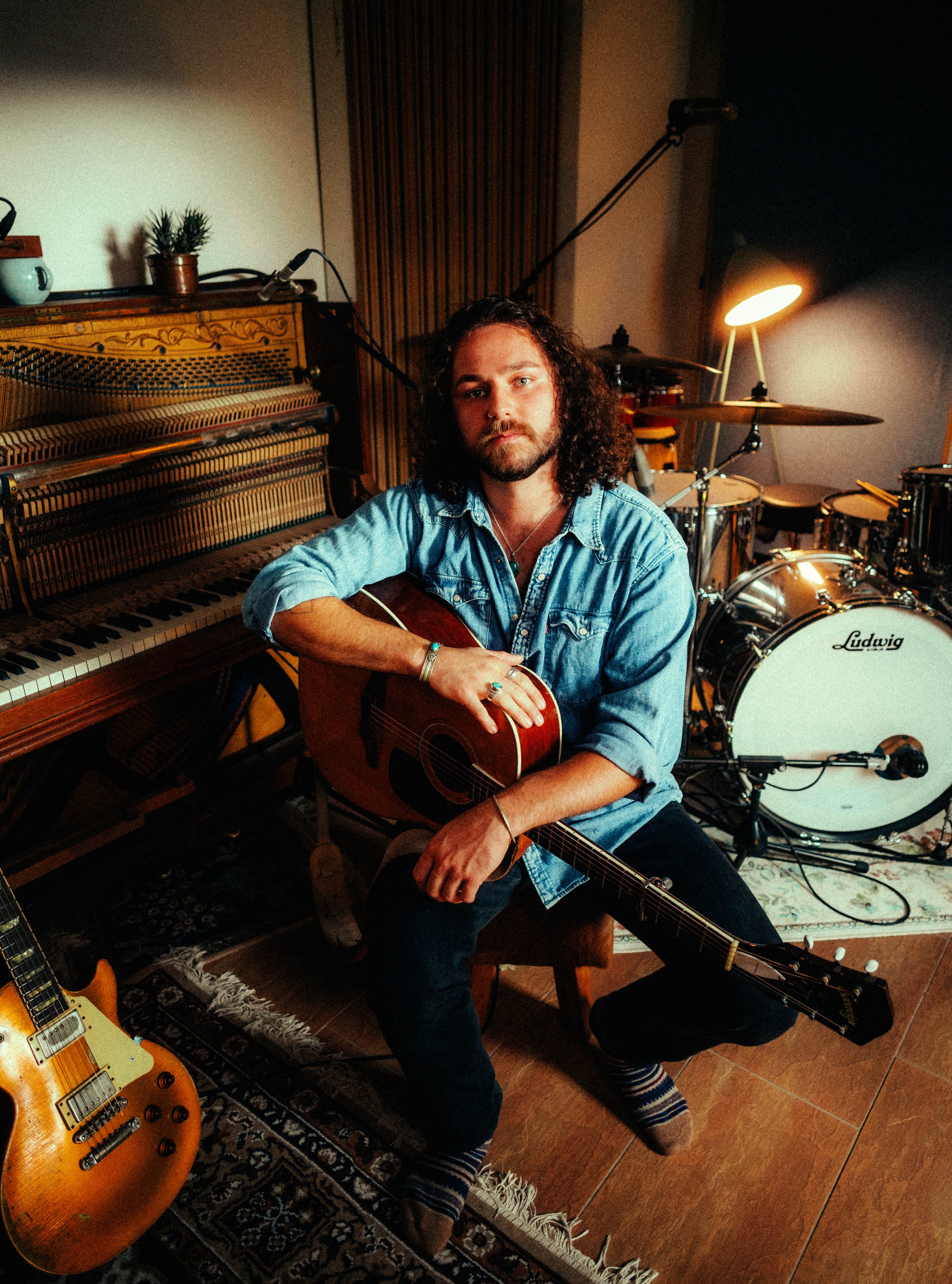 Ein Mann mit lockigem Haar und Bart sitzt mit einem akustischen Gitarreninstrument in einem Musikstudio, umgeben von einem Klavier, einem Schlagzeug und weiteren Musikinstrumenten.