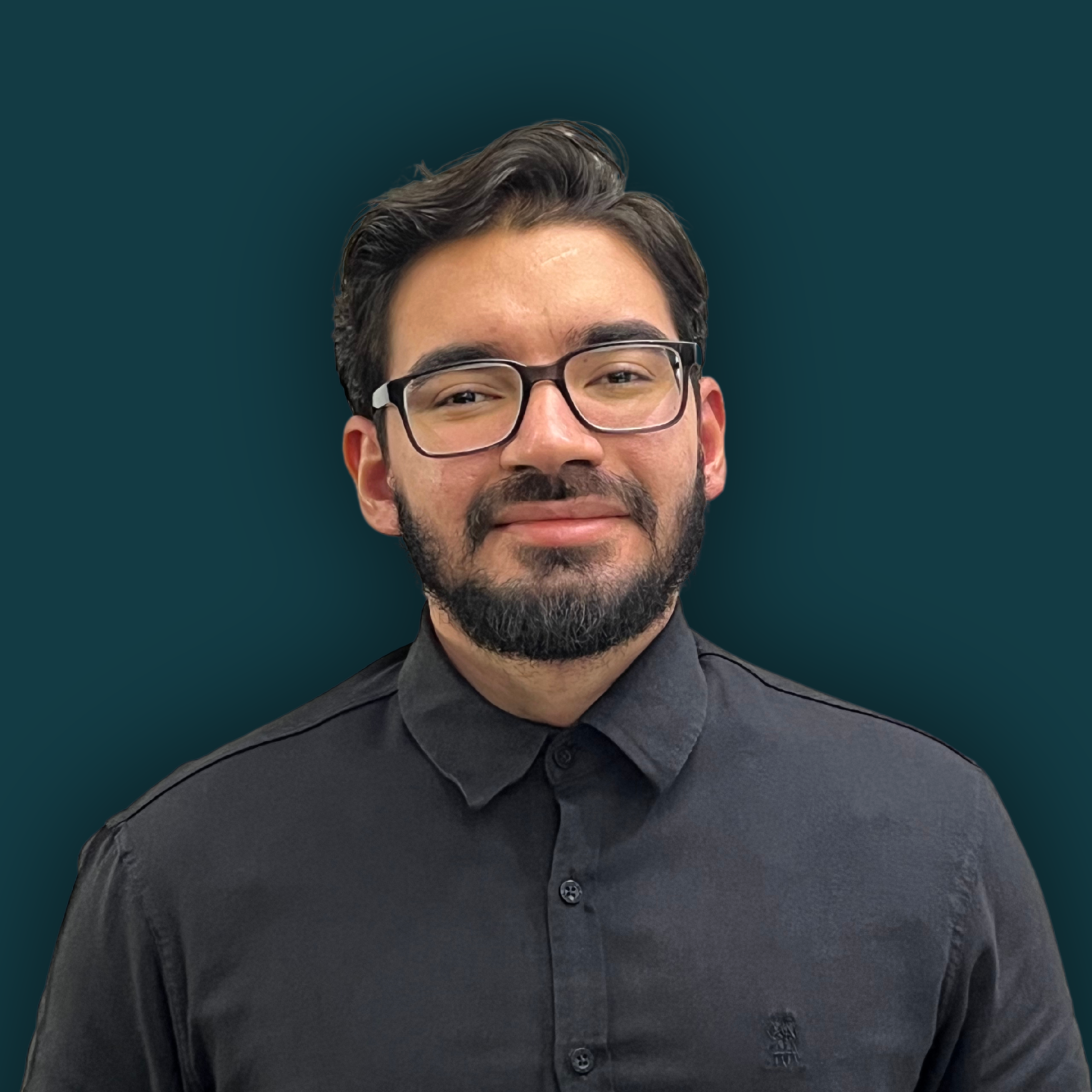 Close-up of a man with dark hair, glasses, and facial hair wearing a dark button-up shirt, smiling against a plain dark teal background.