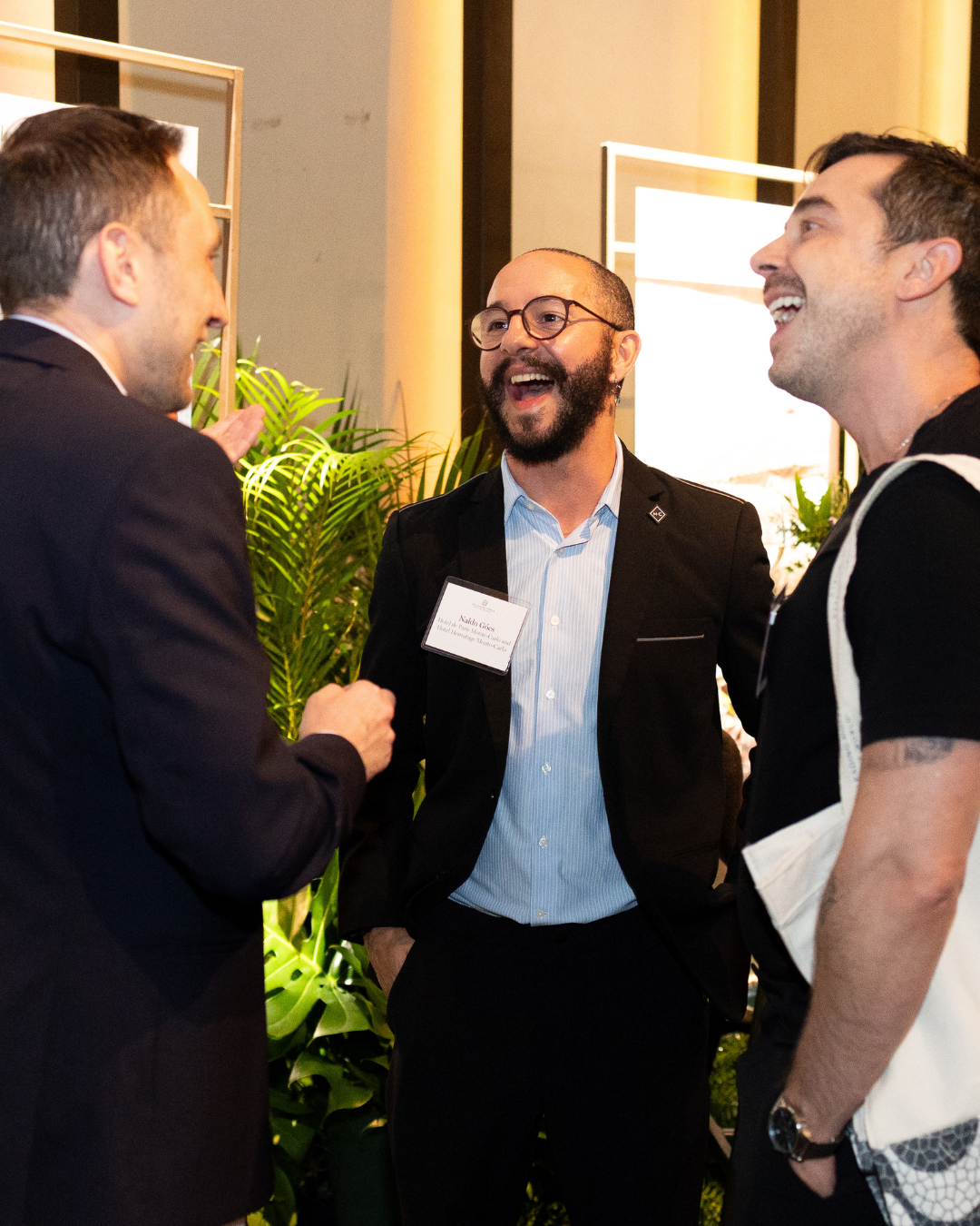 Three men are casually talking and laughing at a social event with plants and bright lights in the background.