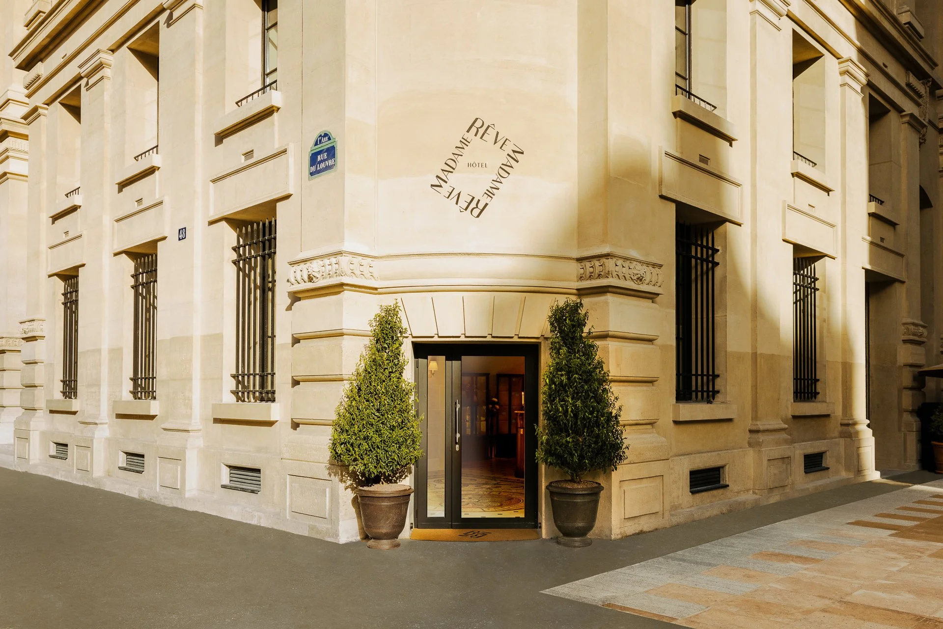 The entrance of a hotel called Madame Rêve, located on Rue du Louvre, with two potted trees on either side of the door, and a building with ornate architectural details and barred windows.