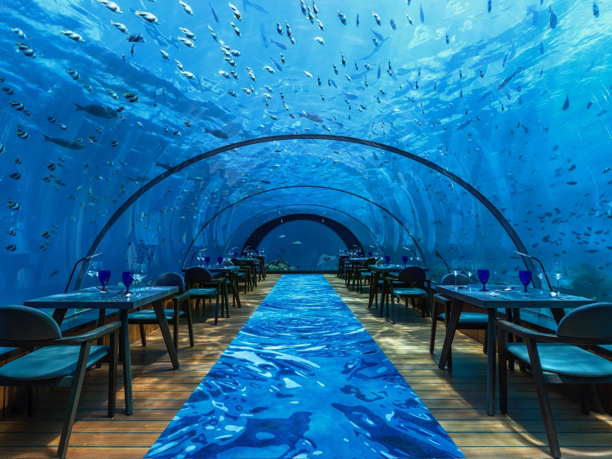 Underwater themed restaurant with tables and chairs along a walkway inside a glass tunnel with a large school of fish swimming overhead.