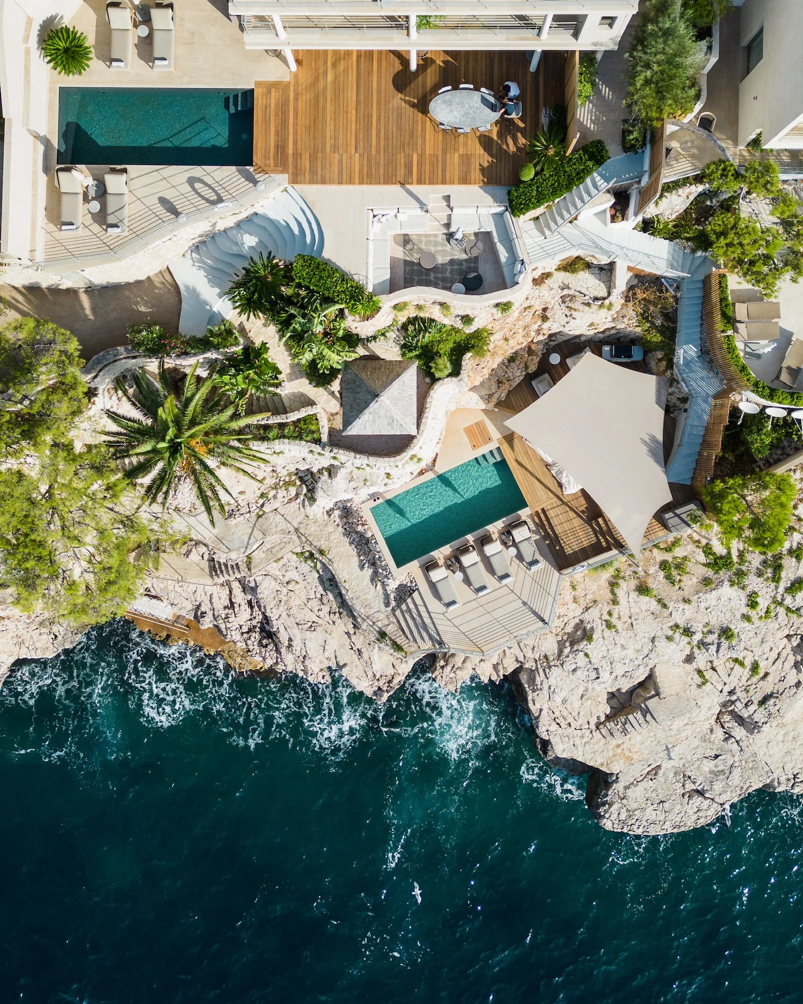 Aerial view of a luxurious coastal property with pools, sun loungers, lush greenery, and a deck, overlooking the ocean.