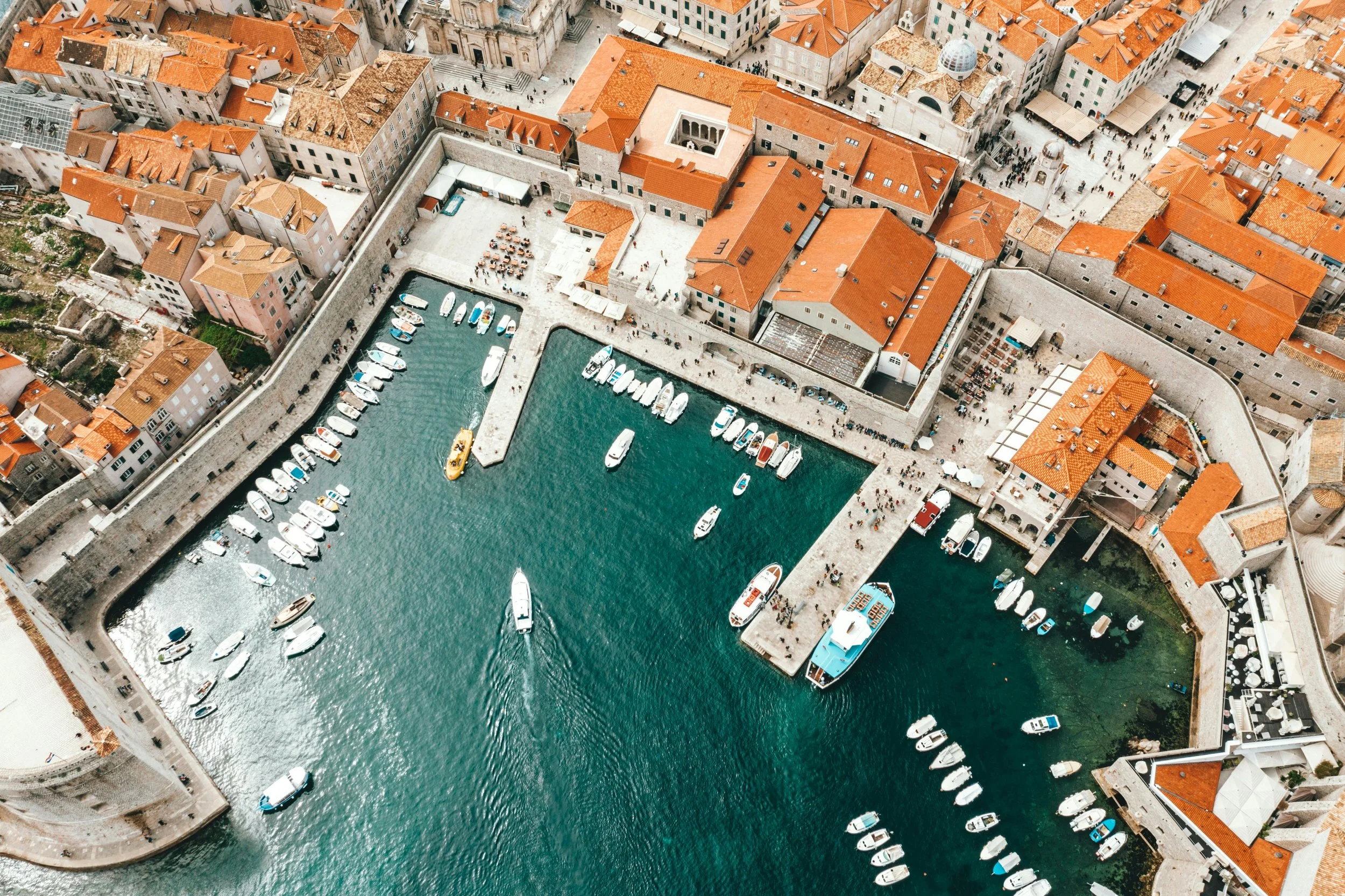An aerial view of a historic harbor with boats docked in a turquoise water, surrounded by old stone buildings and red-tiled roofs, with bustling walkways and city streets.
