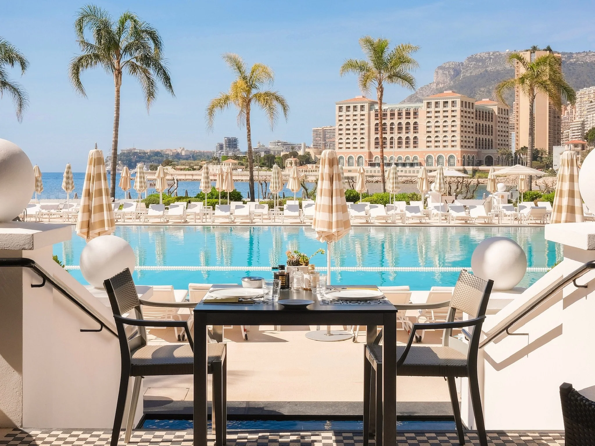 A scenic view of a luxurious outdoor pool area with beige and white striped umbrellas, lounge chairs, and palm trees overlooking the sea with buildings and mountains in the background. There is a dining table with plates and glassware in the foreground.