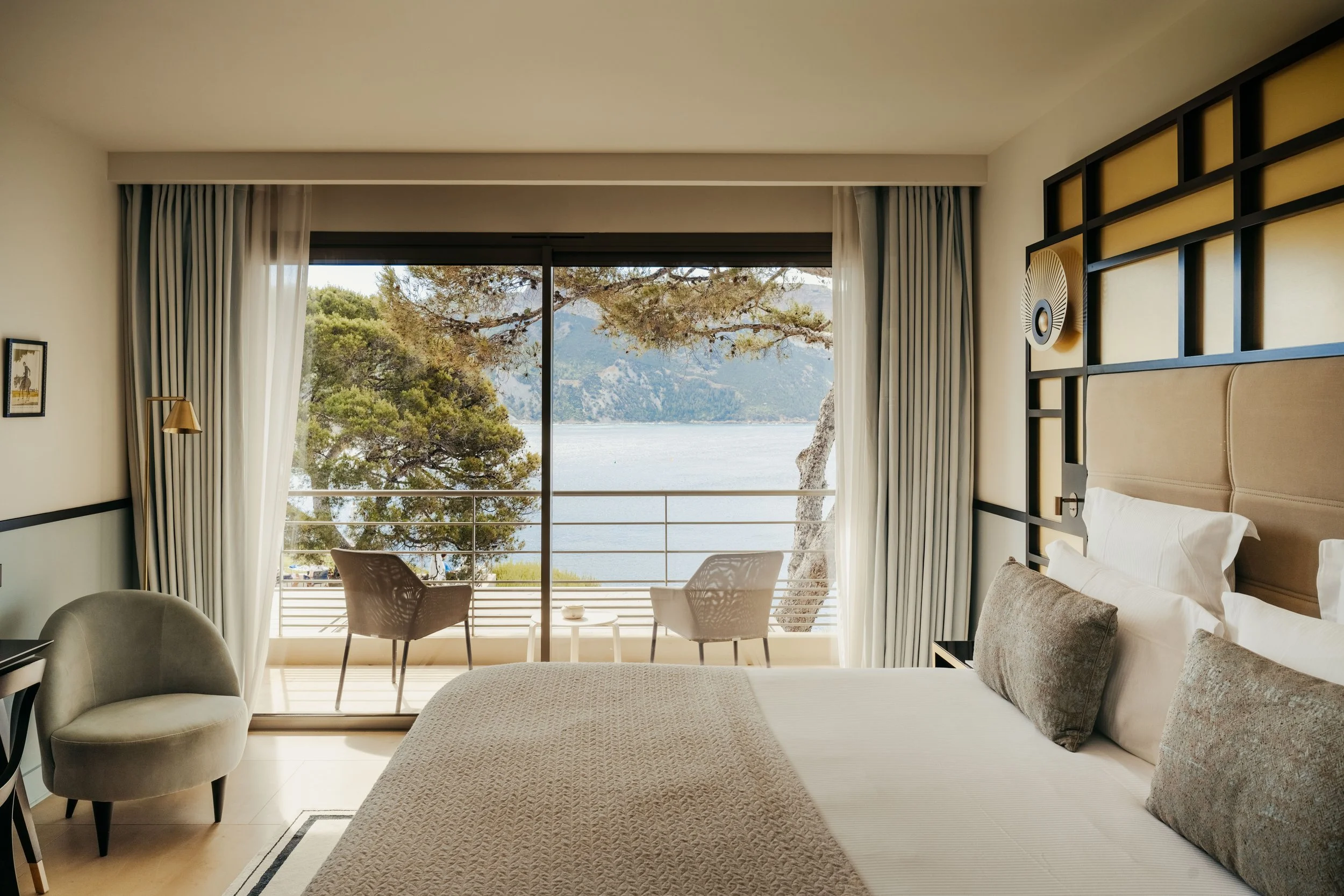 Hotel room with a large bed, beige headboard, pillows, and decorative wall art. Sliding glass doors lead to a balcony with outdoor chairs and a view of trees, water, and mountains.