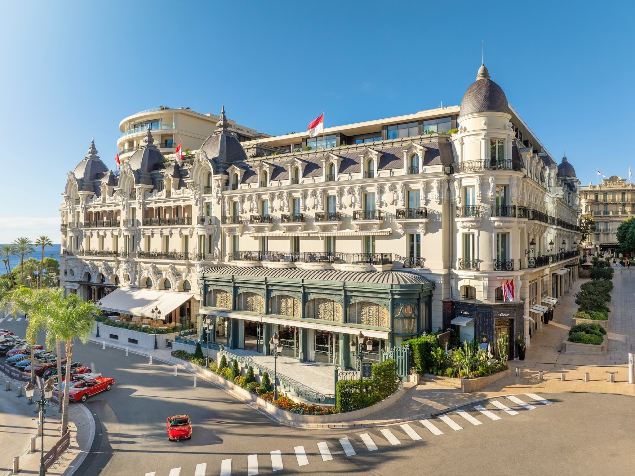 A large, elegant hotel building with ornate architecture, multiple balconies, and conical rooftops. The hotel is situated on a corner with a roundabout and palm trees nearby, under a clear blue sky.