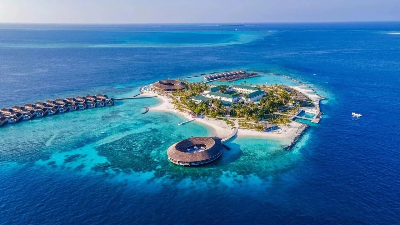 Aerial view of a tropical island resort with overwater bungalows, a circular building, swimming pools, palm trees, sandy beaches, and turquoise ocean waters.