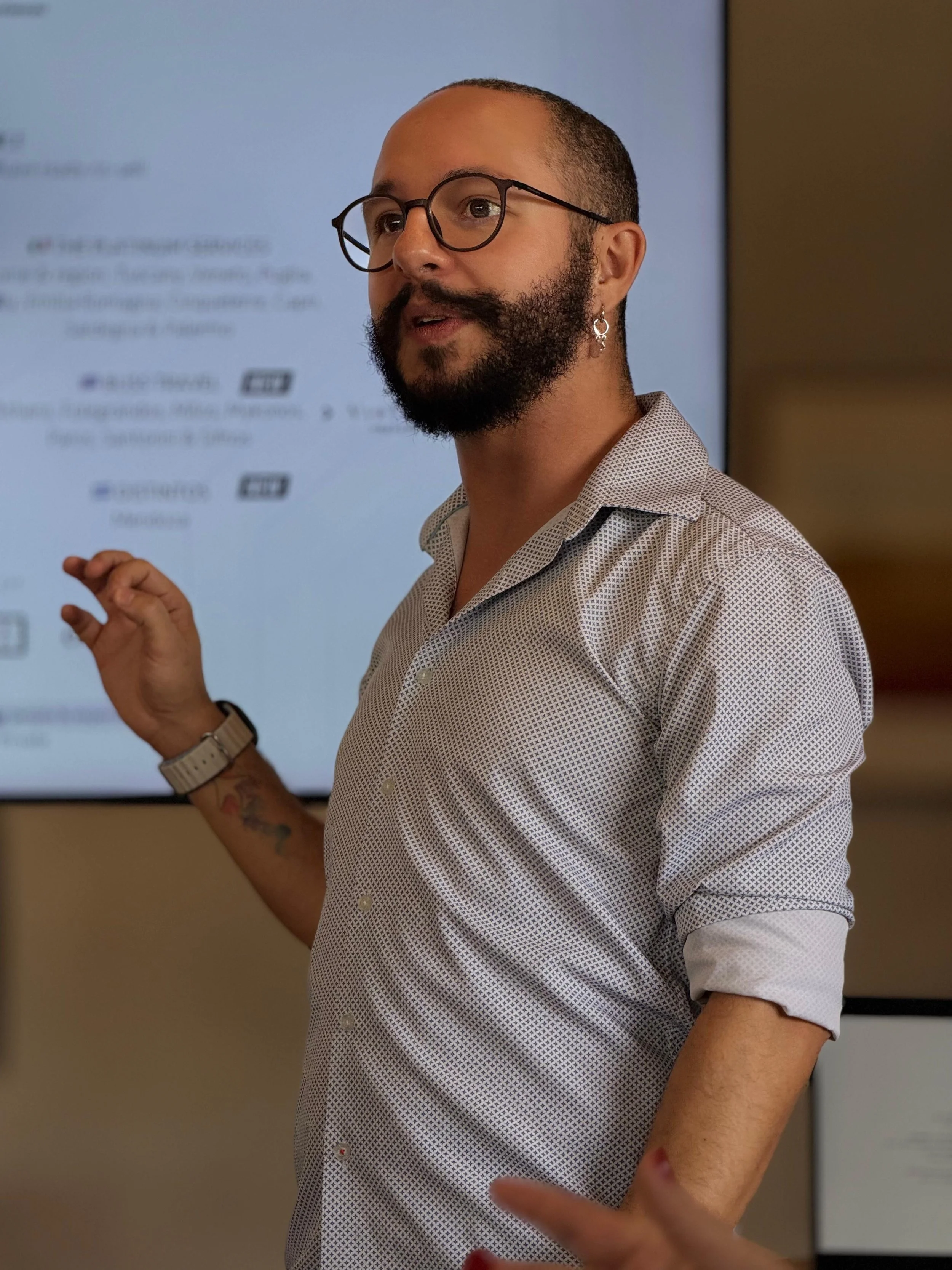 A man with glasses and a beard, wearing a patterned shirt, is giving a presentation in front of a screen with blurred text.
