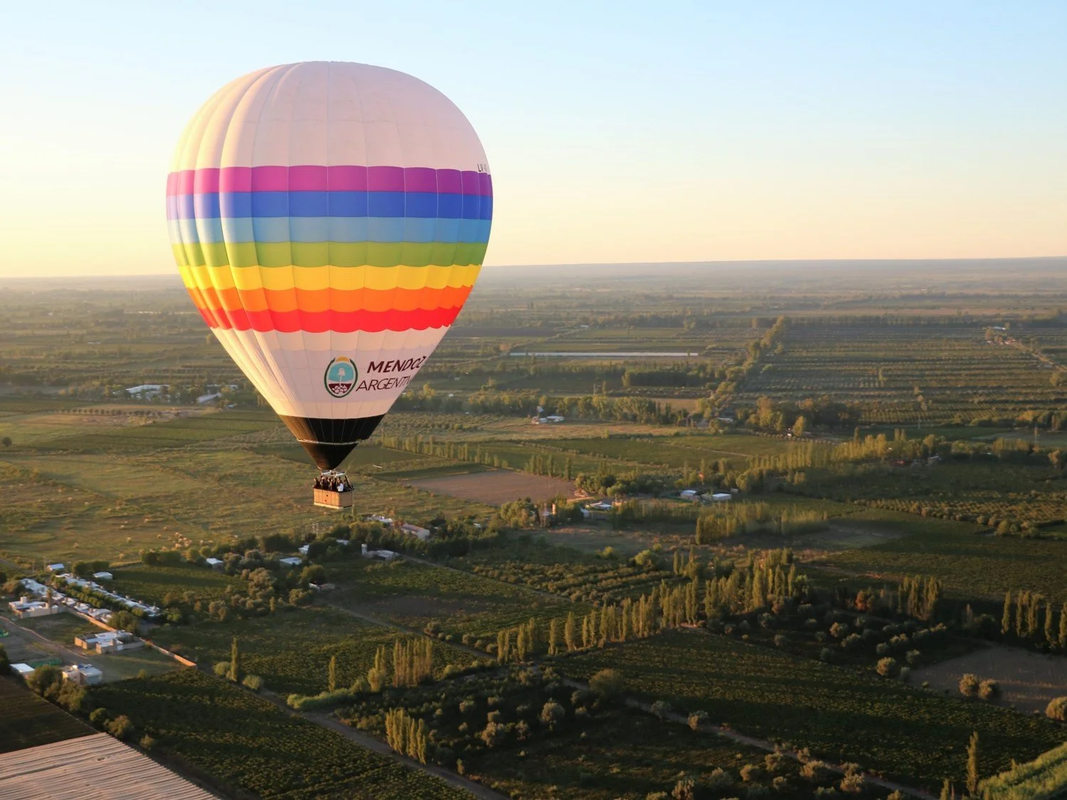 Colorful hot air balloon flying over a green rural landscape at sunrise or sunset, with fields and trees.