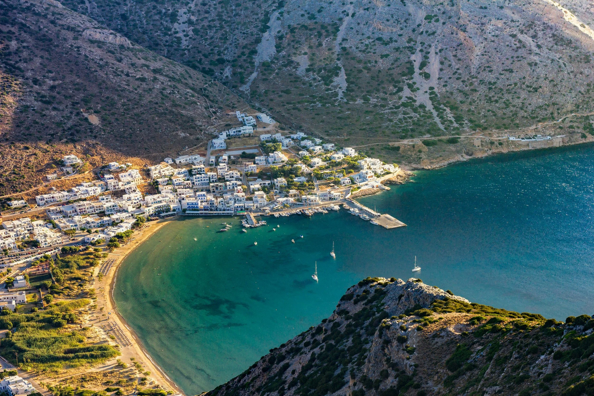 Aerial view of a small coastal village with white buildings, a curved beach, and a harbor with boats, situated between mountains and the sea.