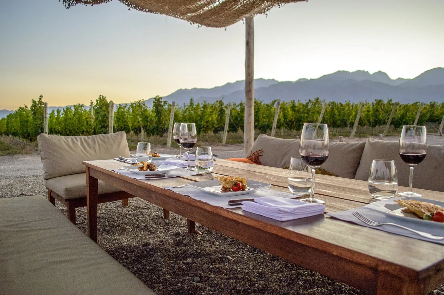 Outdoor dining table set with plates, glasses, and wine in a vineyard with mountains in the background during sunset.