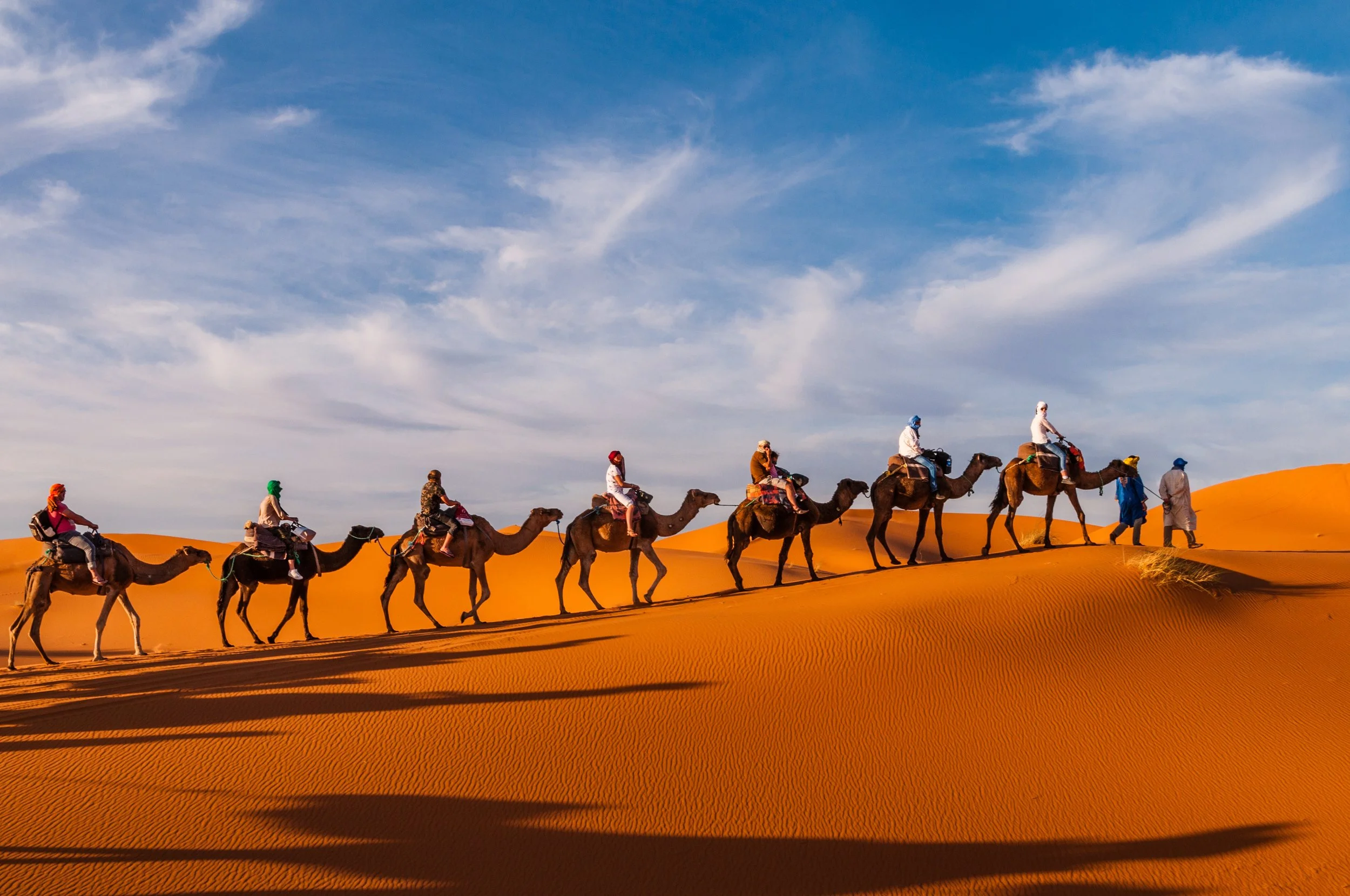Group of people riding camels in desert under blue sky with clouds.