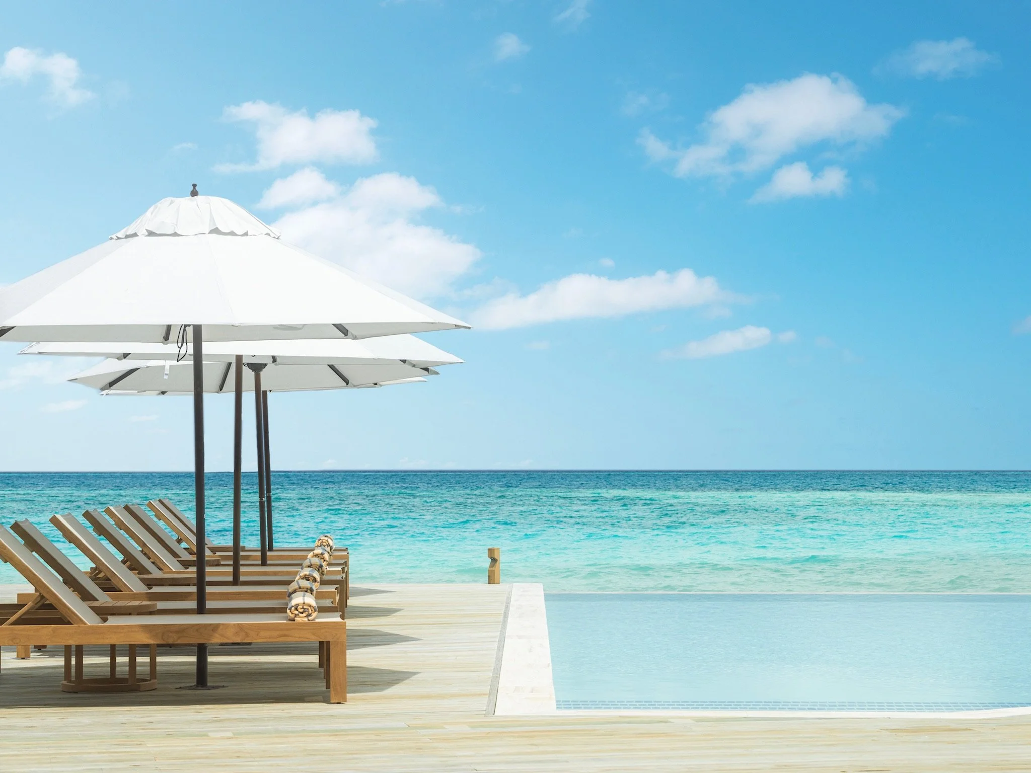 Beach scene with wooden lounge chairs and white umbrellas facing the ocean under a blue sky with scattered clouds.