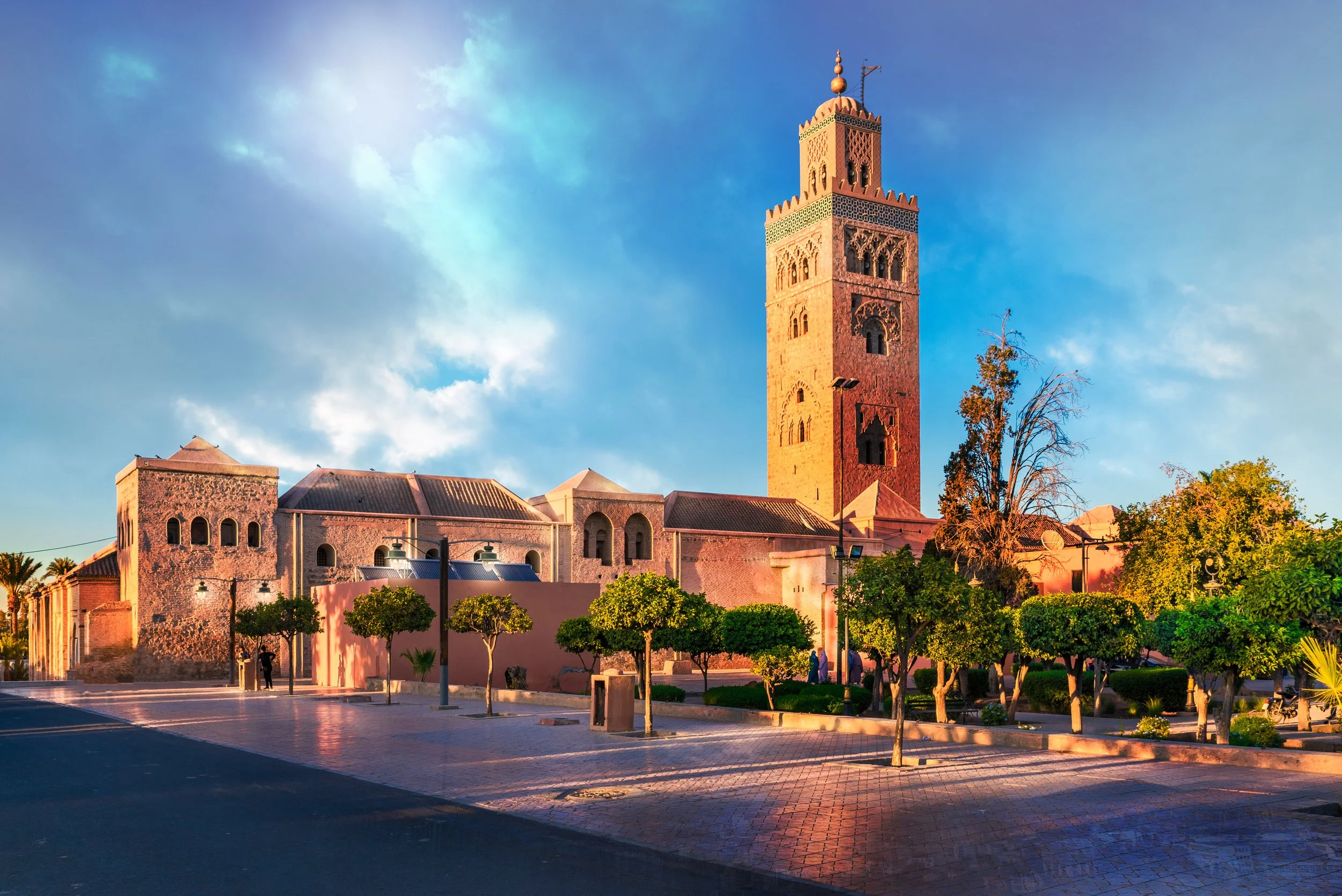 A tall, ornate tower with arched windows and a decorative top, part of a historic building, stands against a bright blue sky with clouds. The foreground features a paved area with small, green, demarcated trees and a few streetlights.