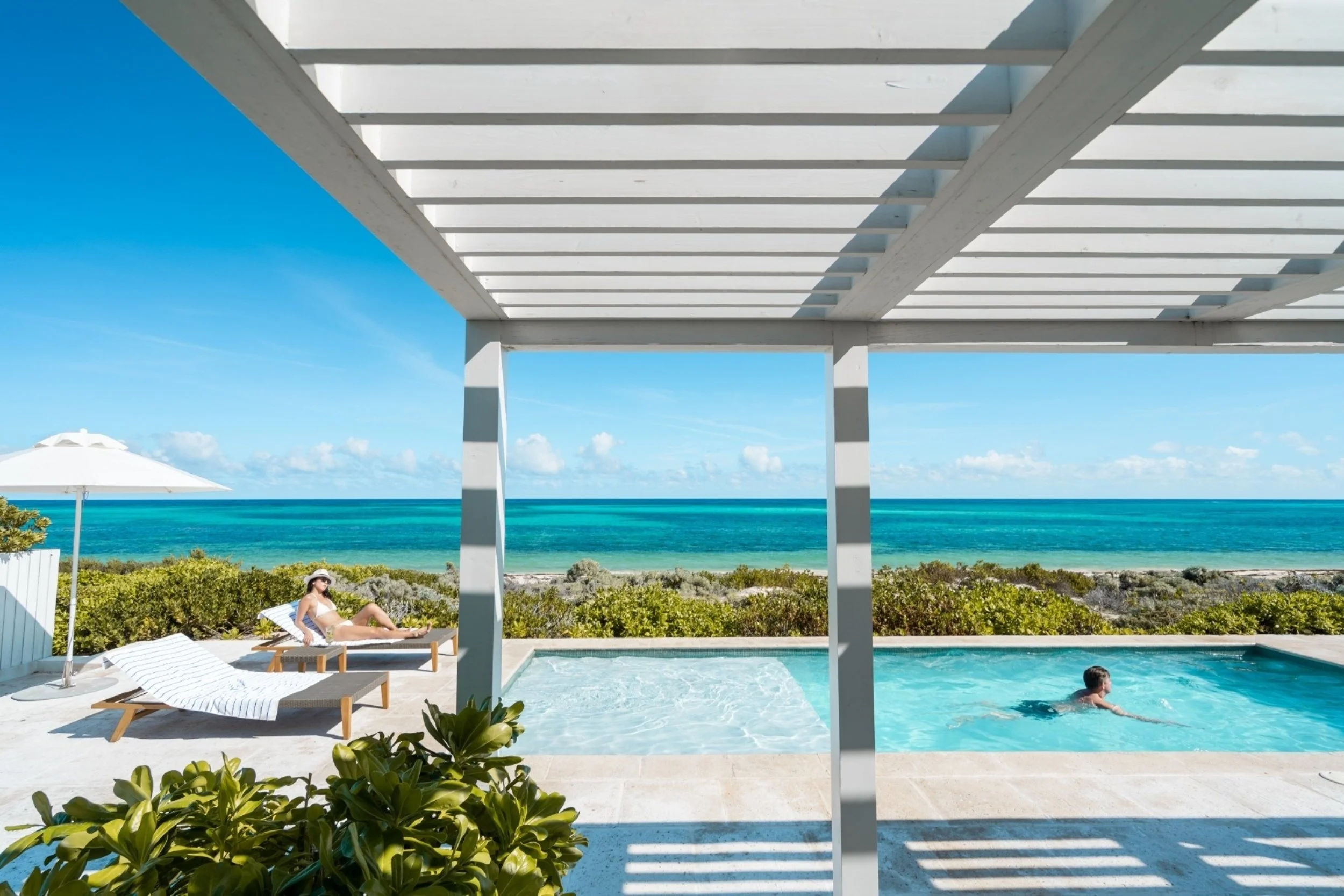 A woman in a white swimsuit and hat relaxing on a lounge chair by a swimming pool at a beachside location. There are umbrellas, green bushes, and the ocean in the background under a partly cloudy sky.